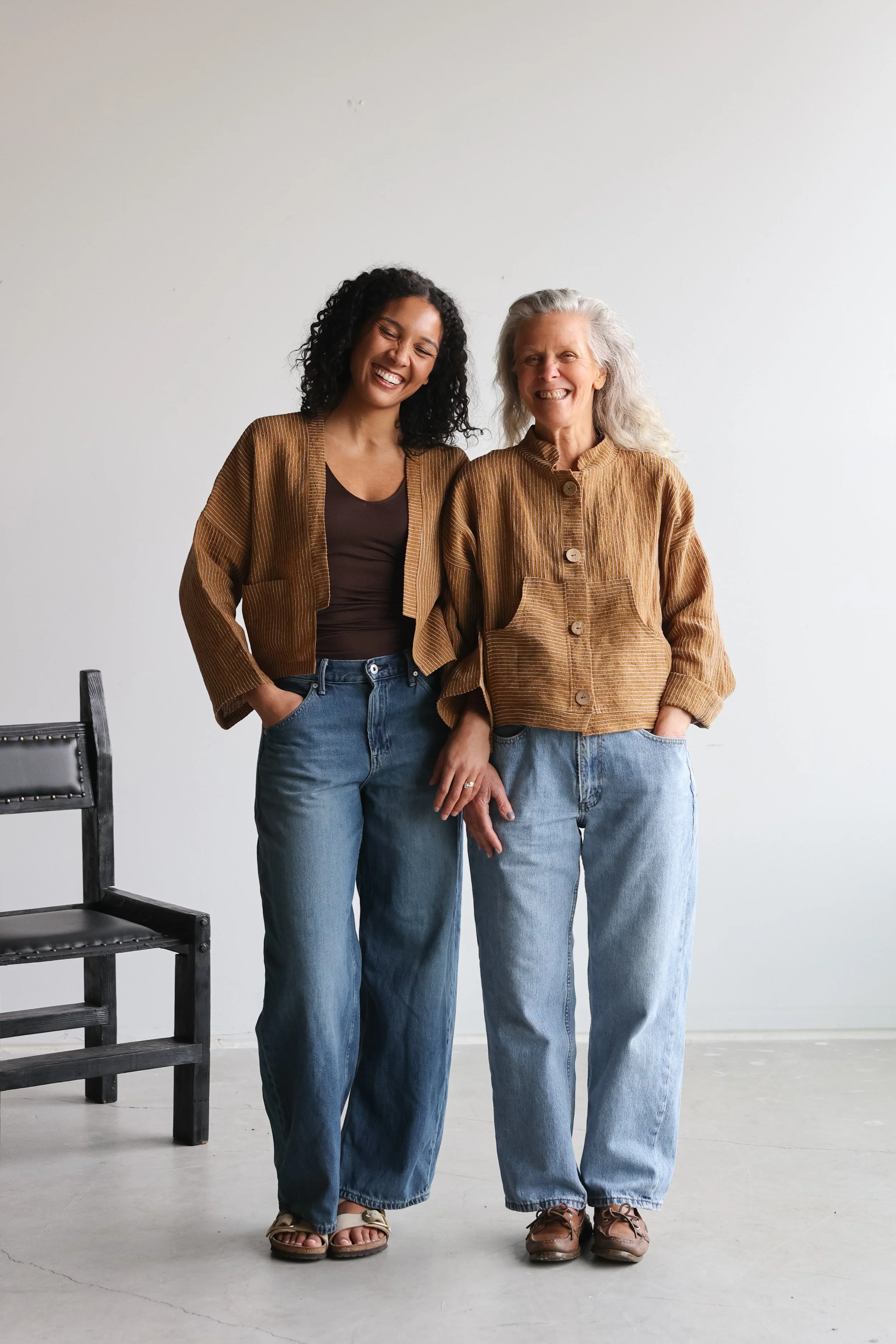 Two women standing close together, smiling, in a plain white room with a black chair partially visible to the left.