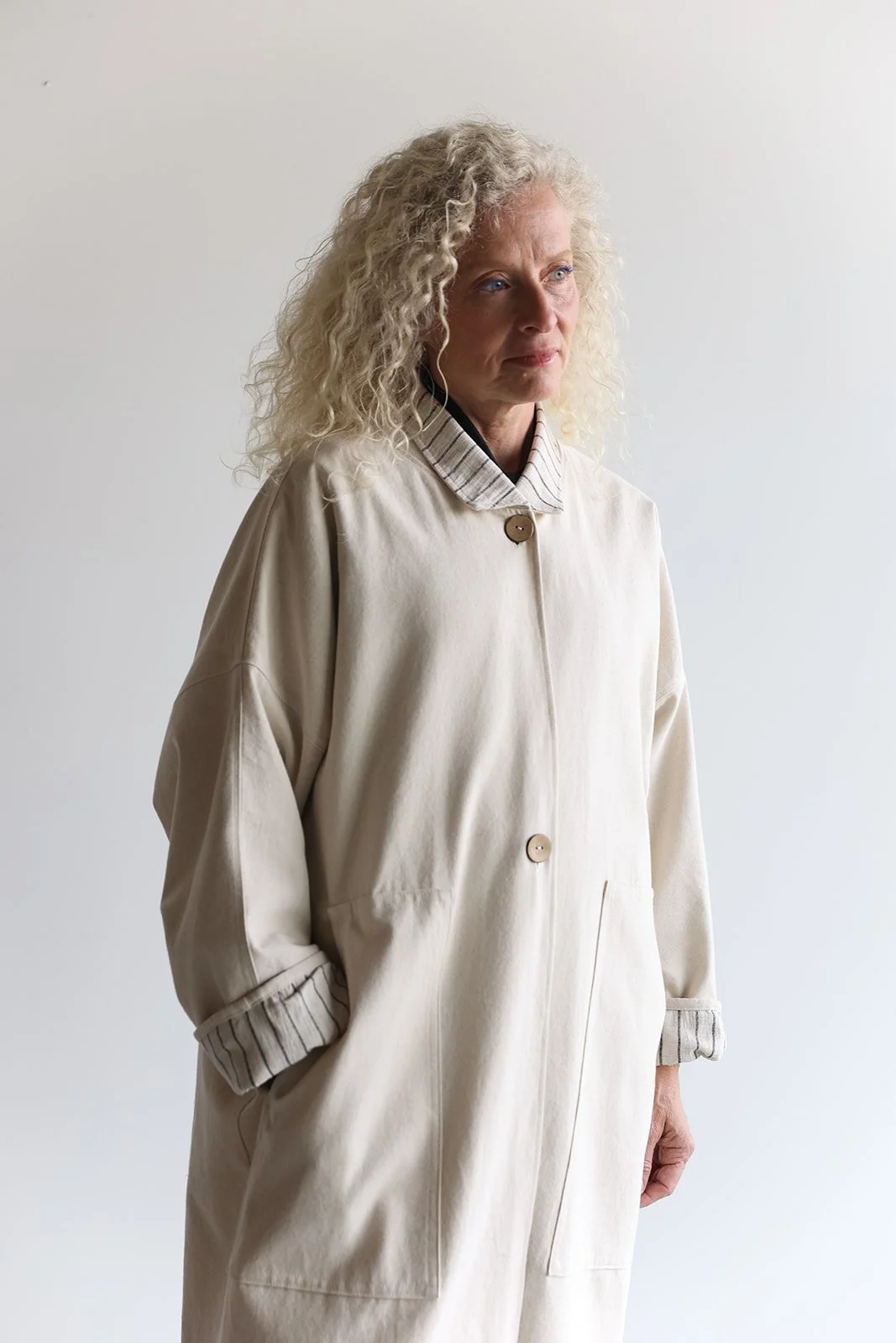 Front view of a woman wearing a canvas linen trench coat in Sand looking right with left hand in pocket