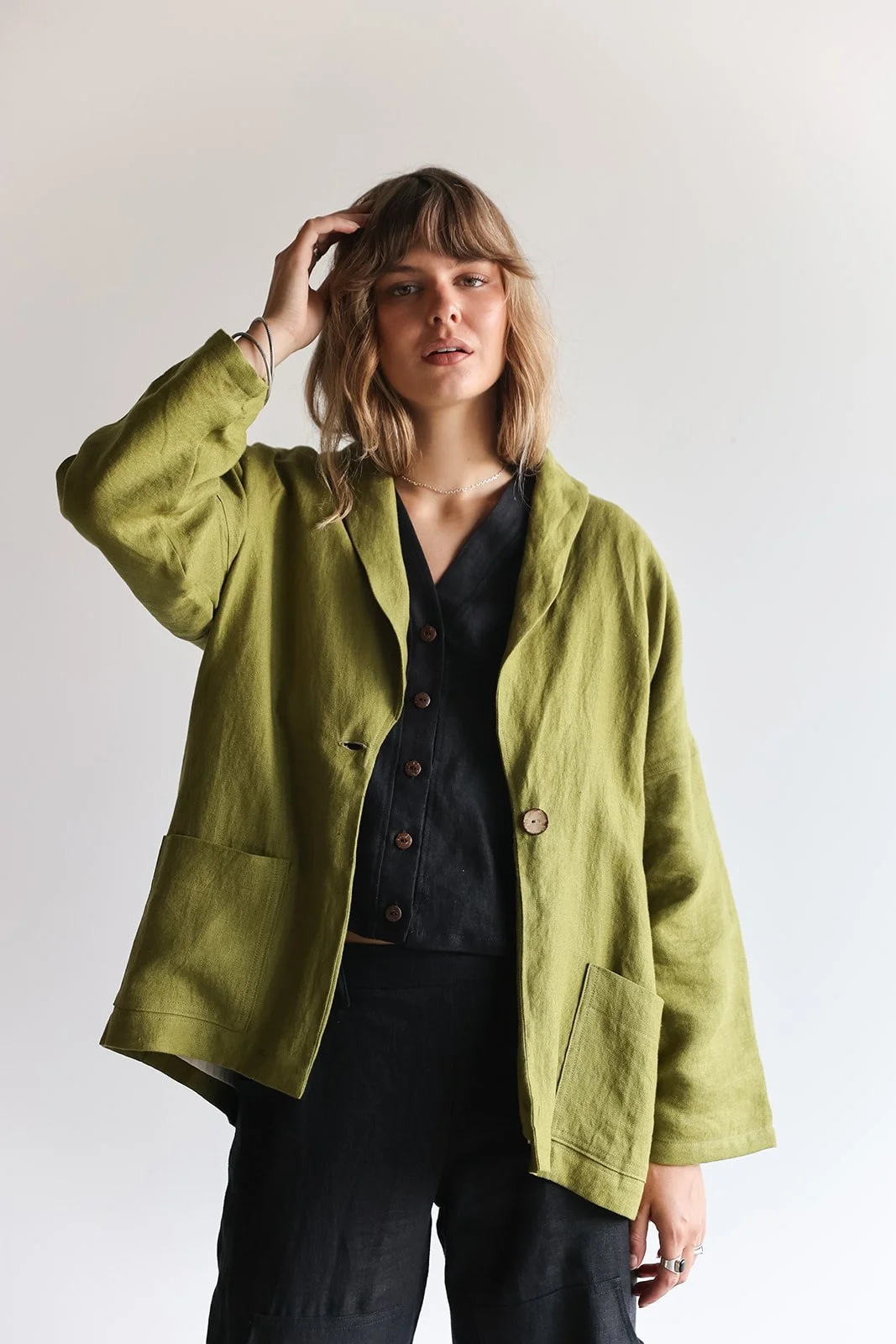 Front view of a woman wearing a linen blazer in Pesto adjusting hair with hand