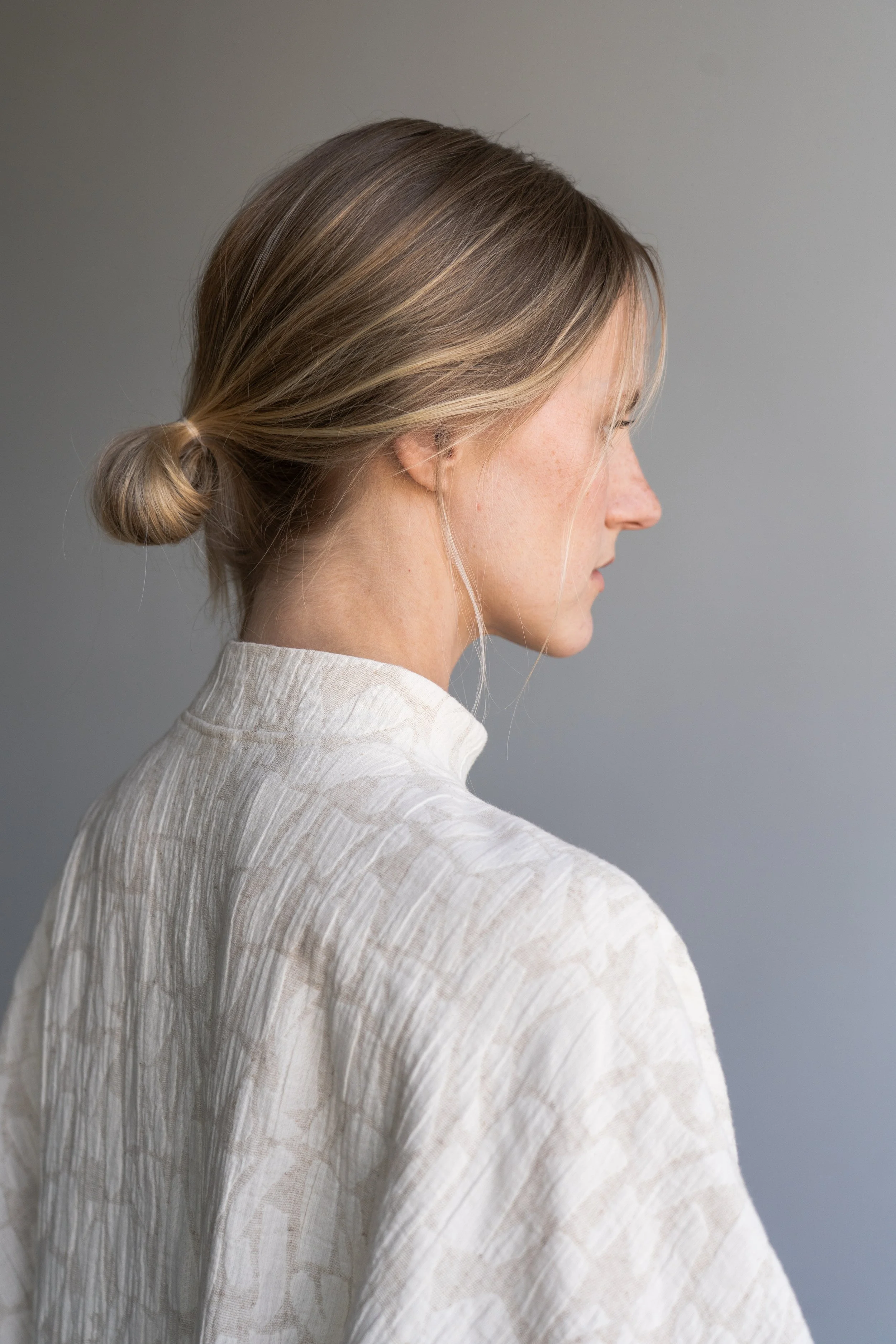 A woman with blonde hair in a low, loose bun, wearing a beige, textured, high-neck top, facing to the right against a gray background.