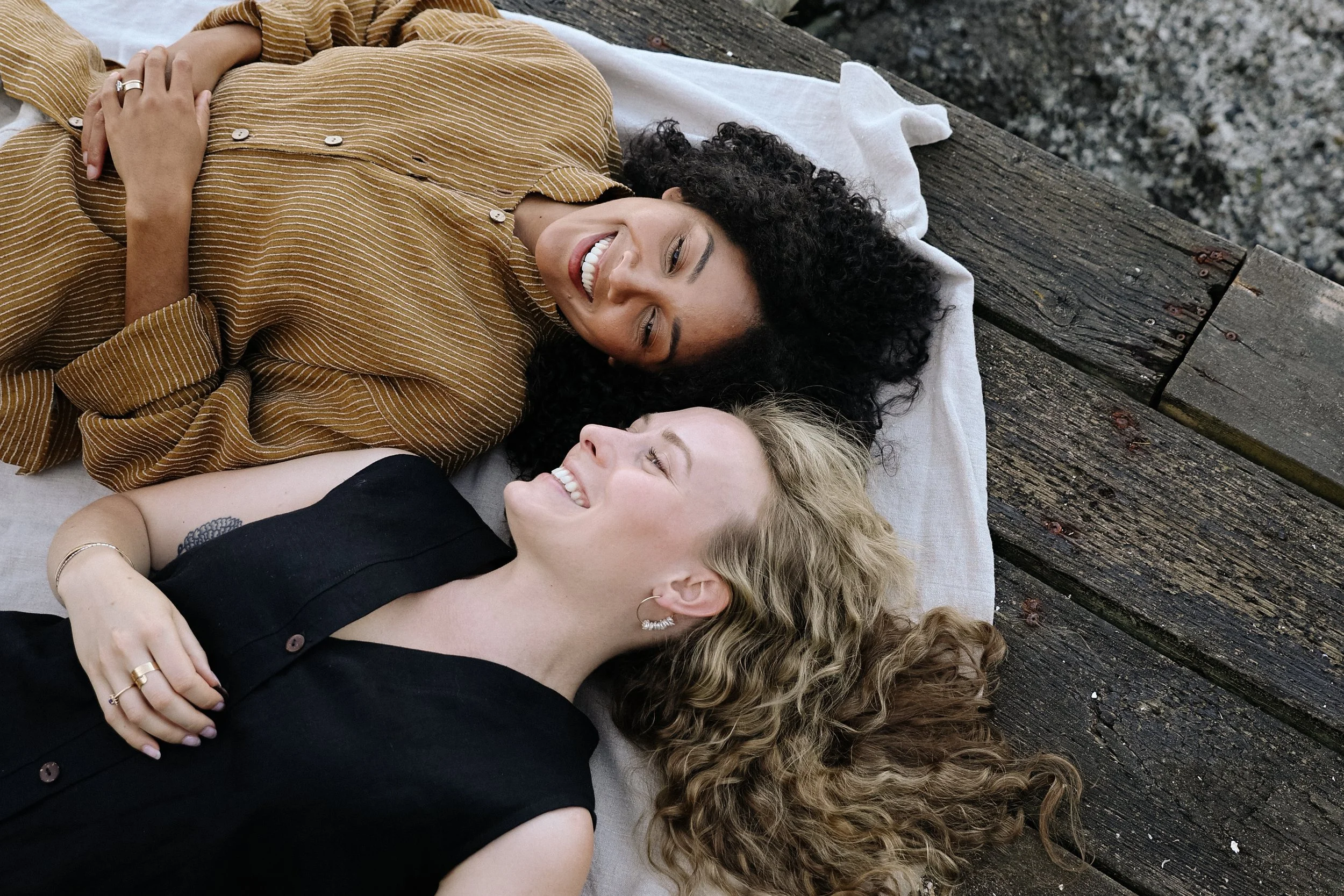 Two women lying on a wooden dock with a gray pillow, smiling at each other, one with curly black hair and the other with long blonde hair.