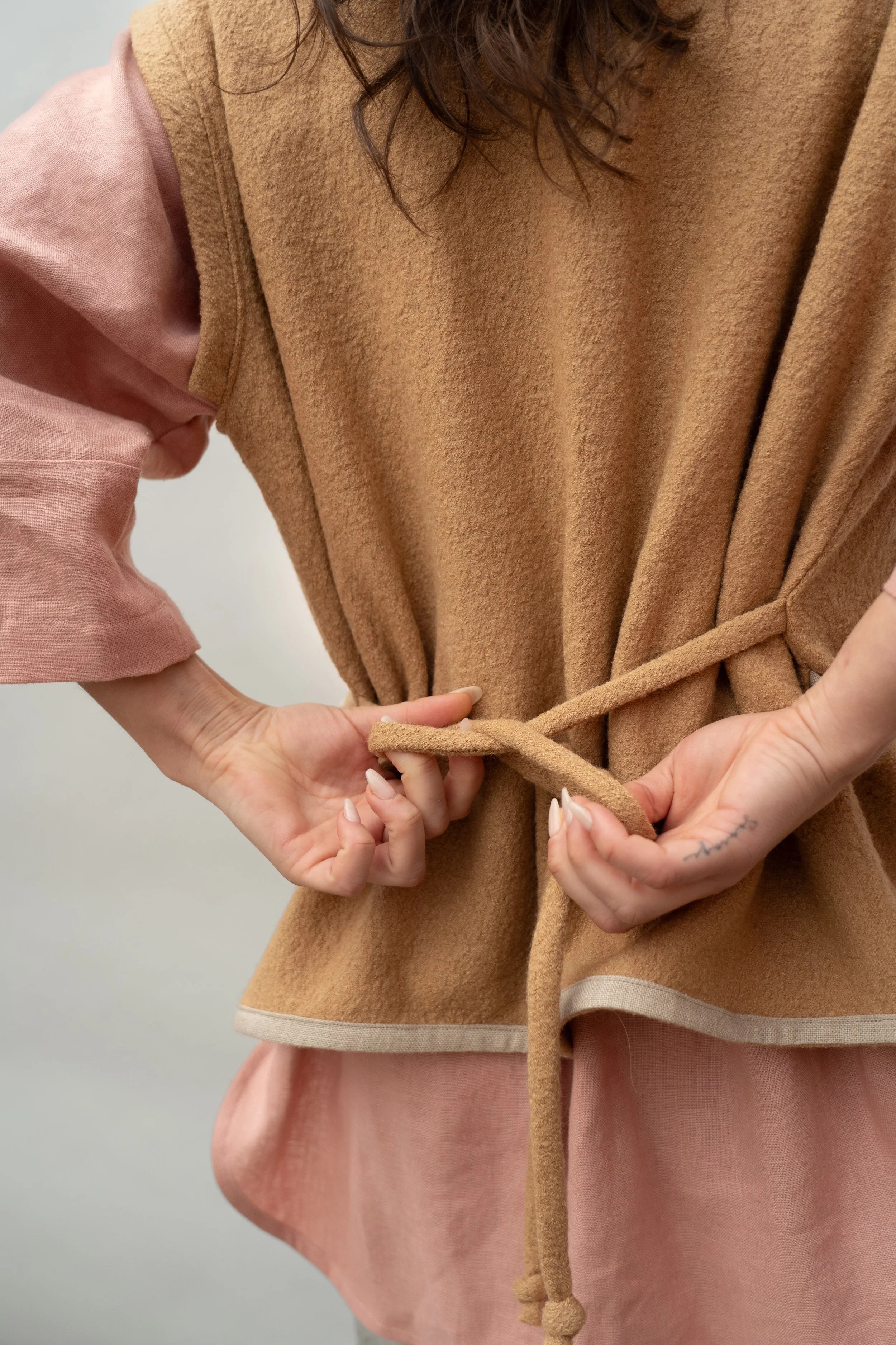 Person tying a beige belt or sash on a brown vest, with pink sleeves and a pink skirt.