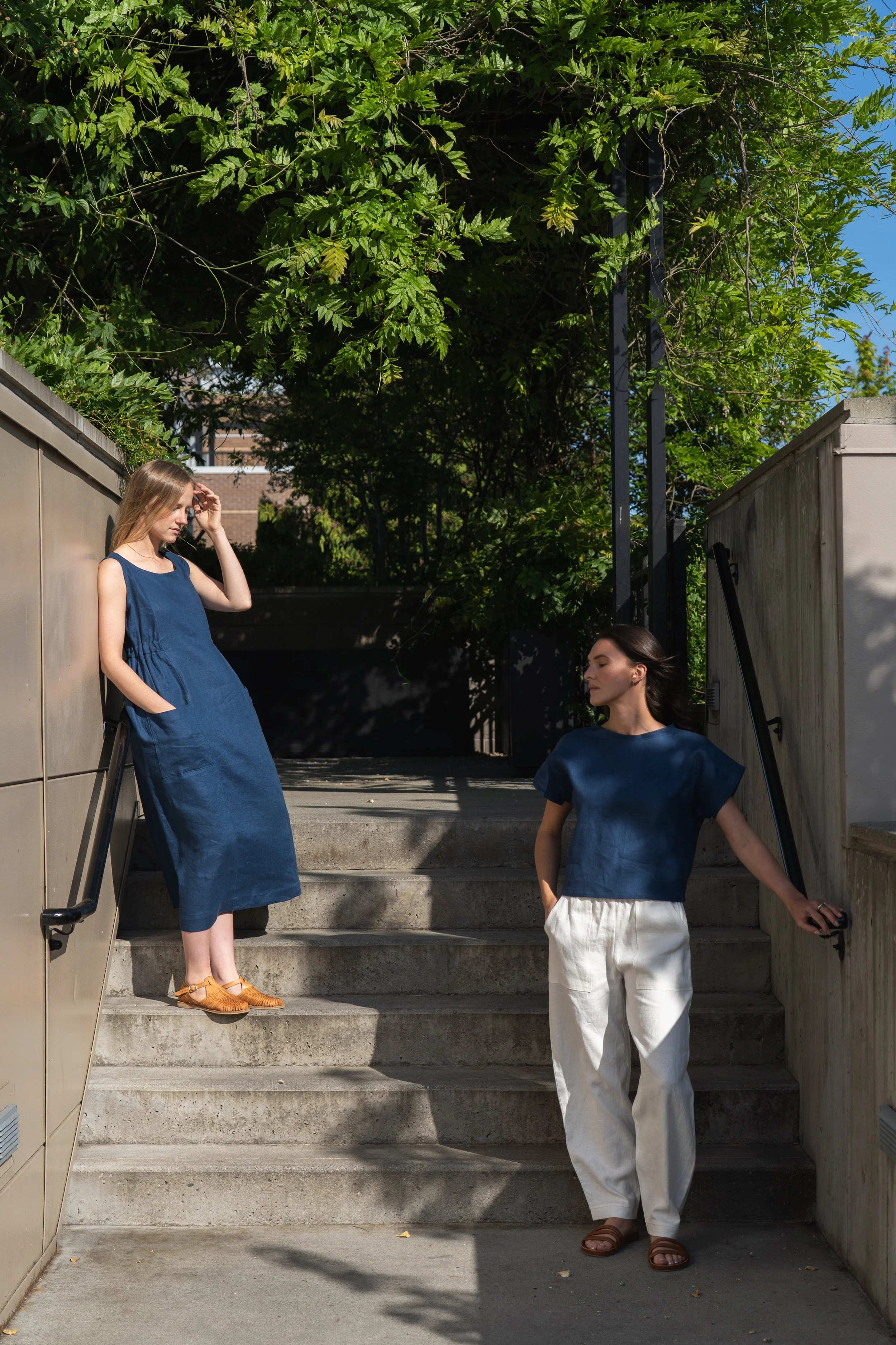 Two women in blue tops, one in a blue dress and the other in a blue shirt and white pants, standing on steps outdoors surrounded by greenery and shadows.