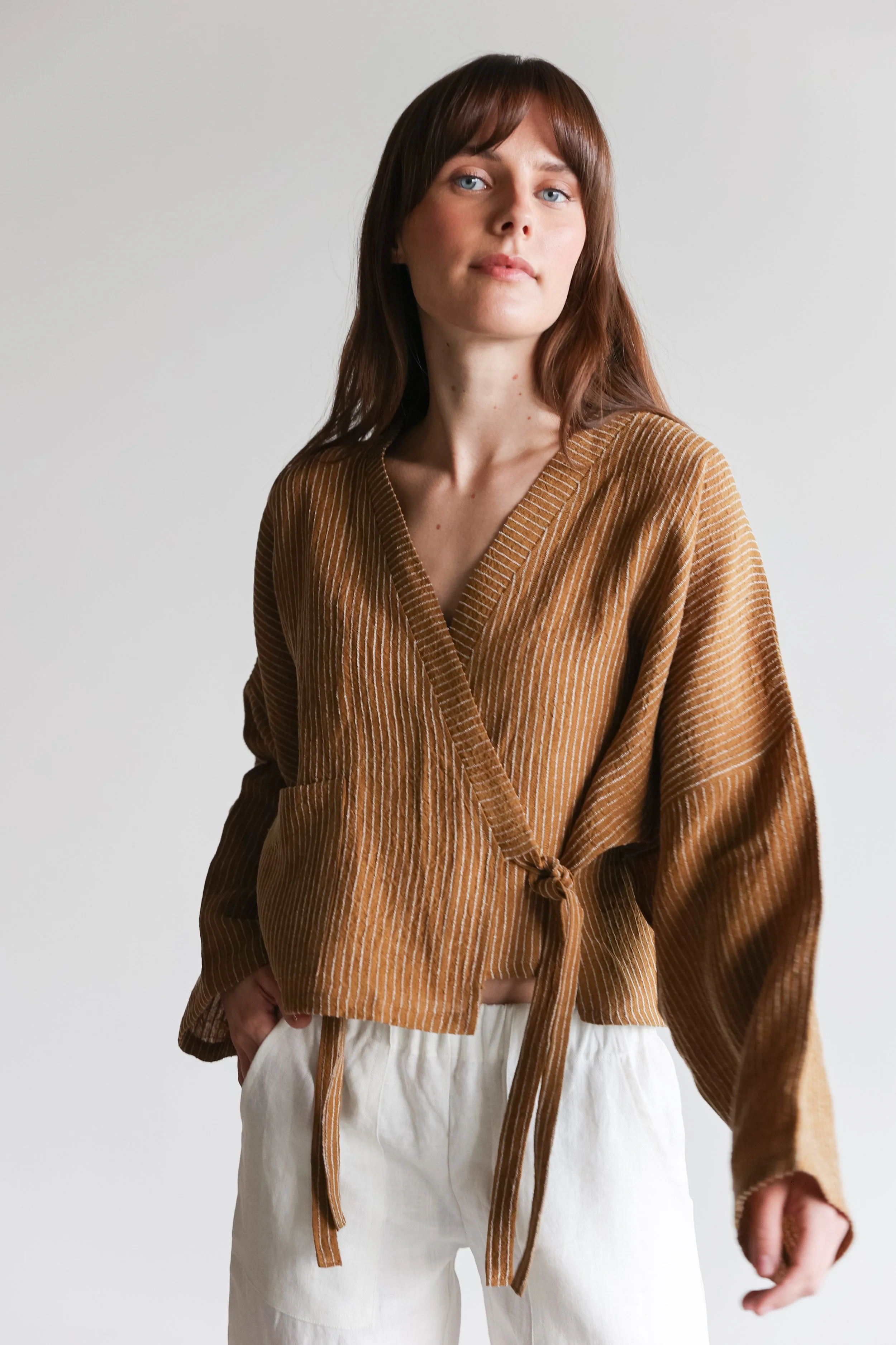 A woman with brown hair and blue eyes wearing a brown striped wrap top with long sleeves and beige pants, standing against a plain white background.