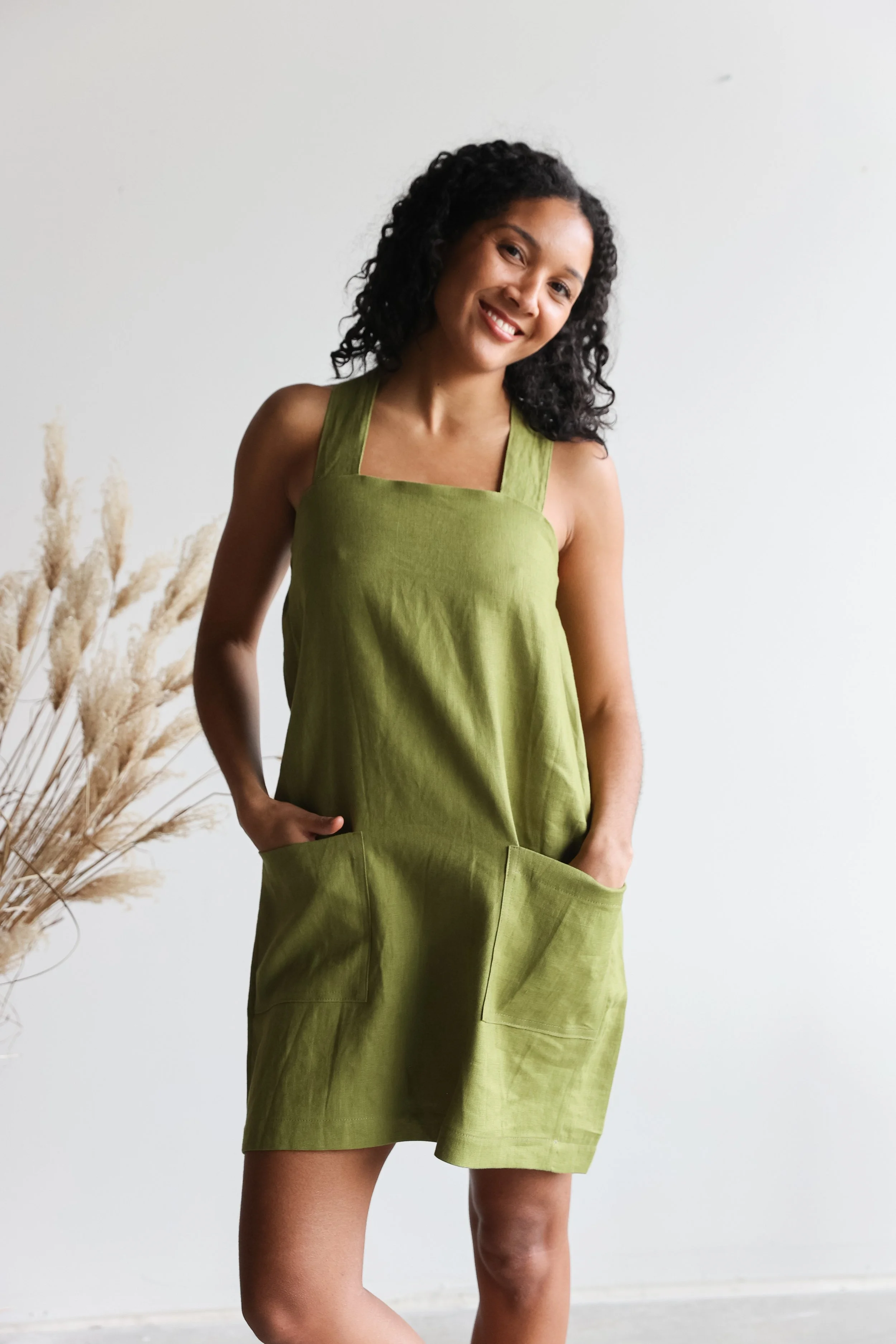 A woman with curly black hair wearing a sleeveless green top and matching green shorts, standing with one hand in her pocket, smiling against a plain white wall with some dried plants on her left.