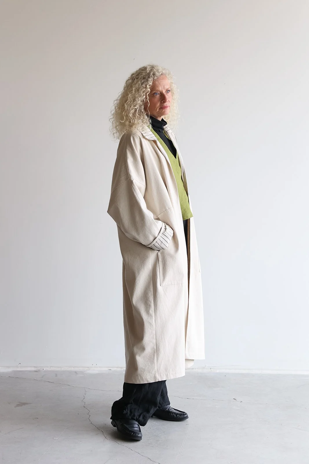 Side view of a woman wearing a canvas linen trench coat in Sand with hands in pockets facing right