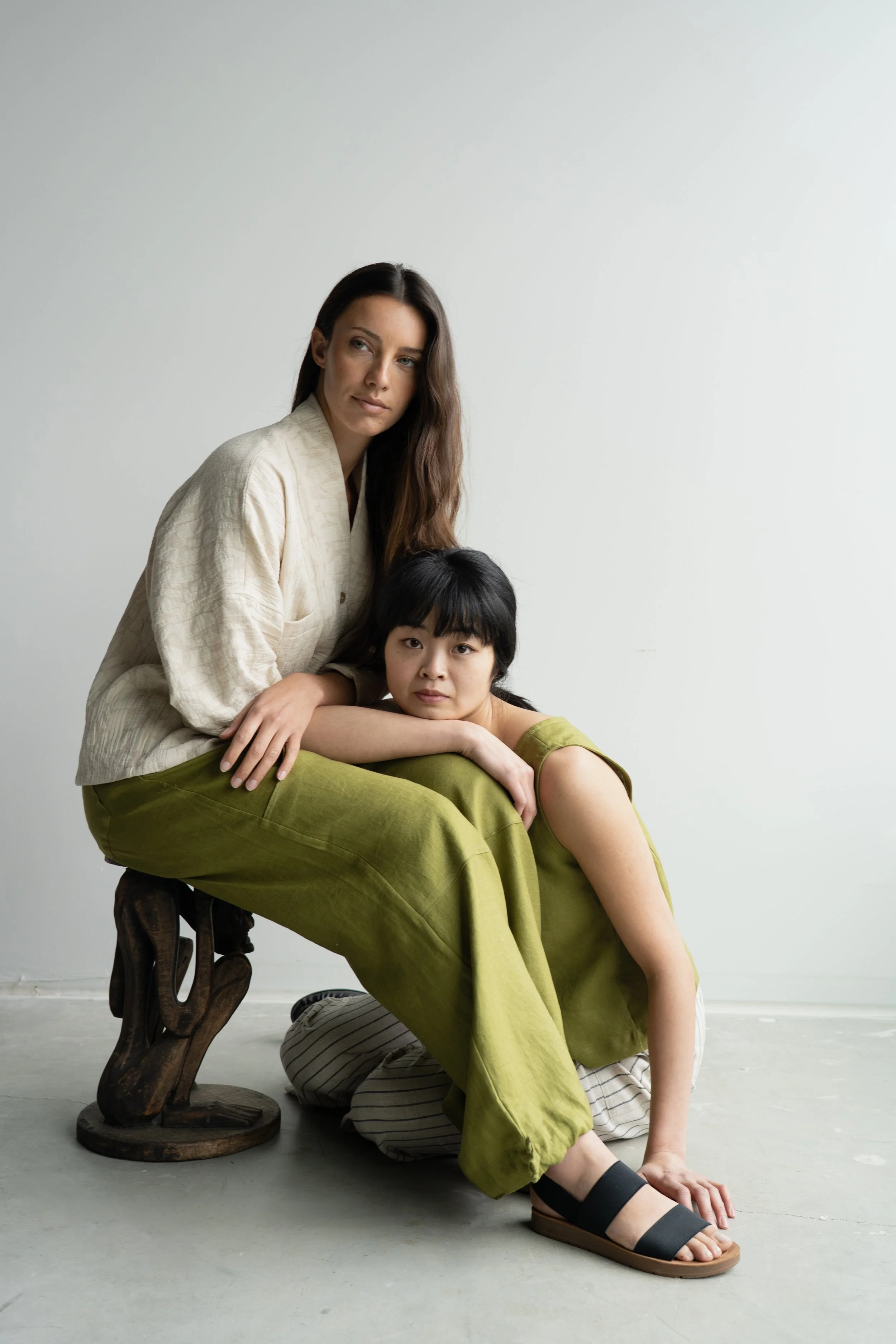 Two women, one with long brown hair and the other with short black hair, sitting on a stool and floor against a plain white wall, with the shorter woman resting her head on the other's lap.