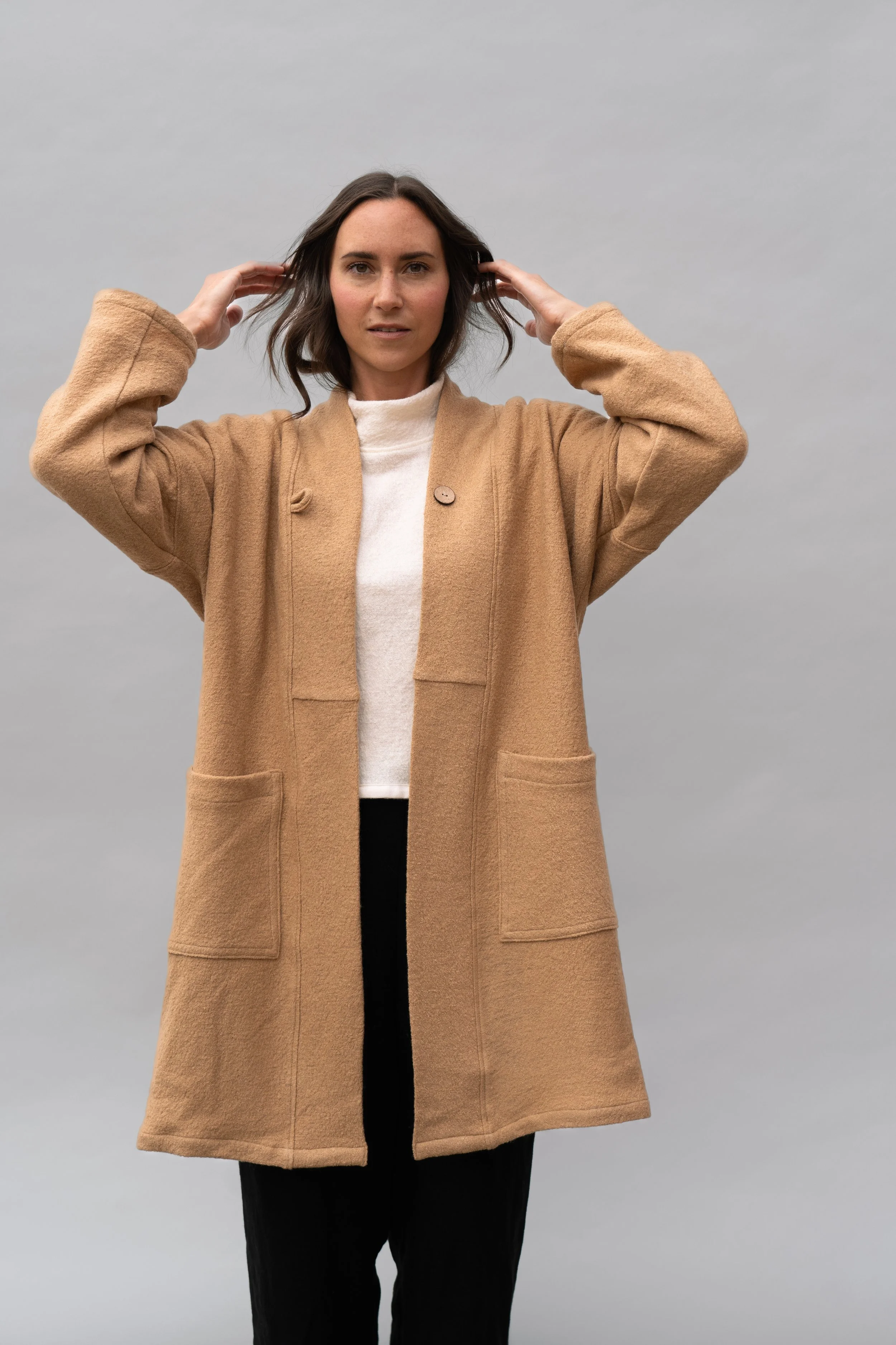 Woman in tan coat standing against a gray background, adjusting her hair.