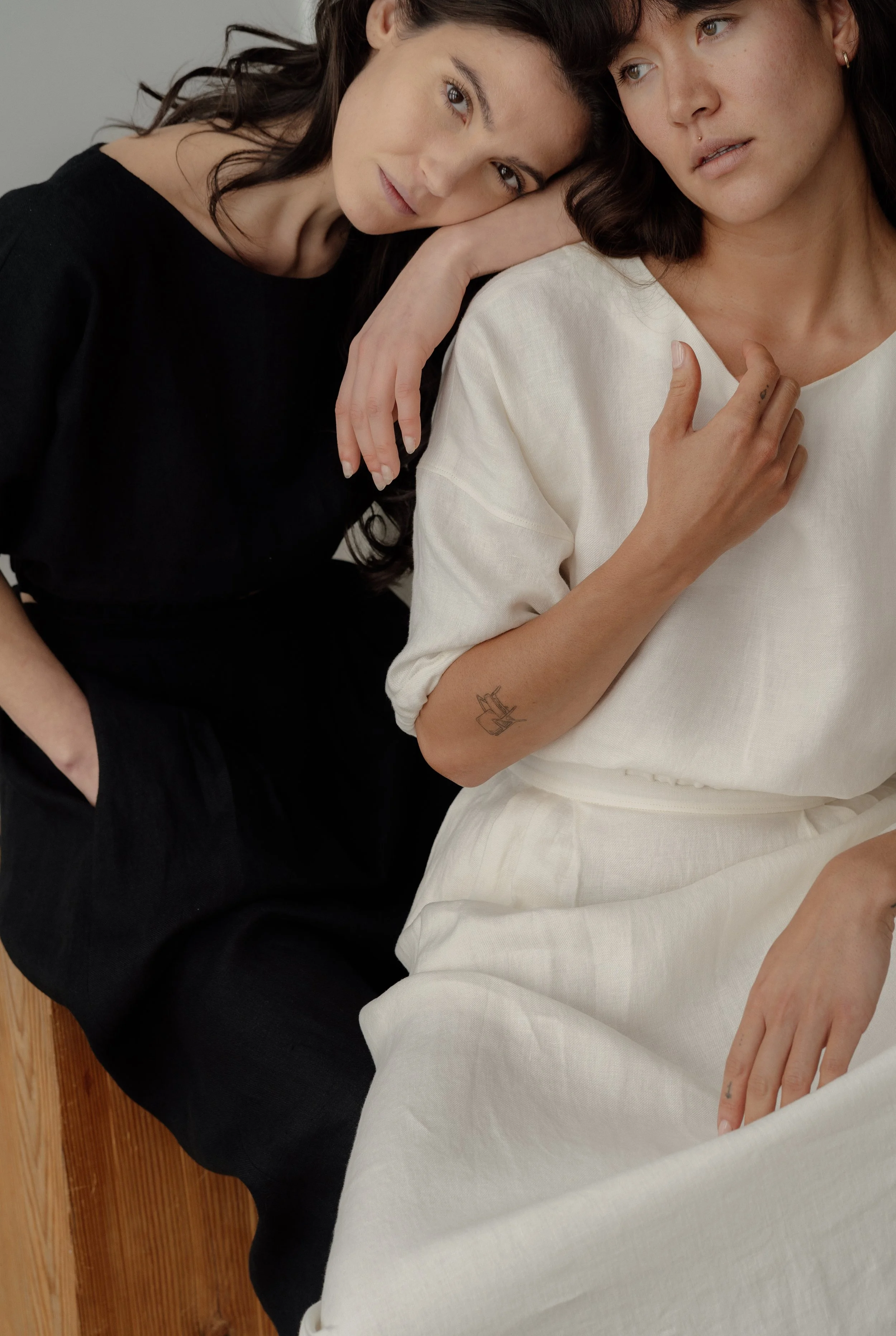 Two women sitting close together, one resting her head on the other's shoulder, both wearing black and white linen outfits, with a simple neutral background.