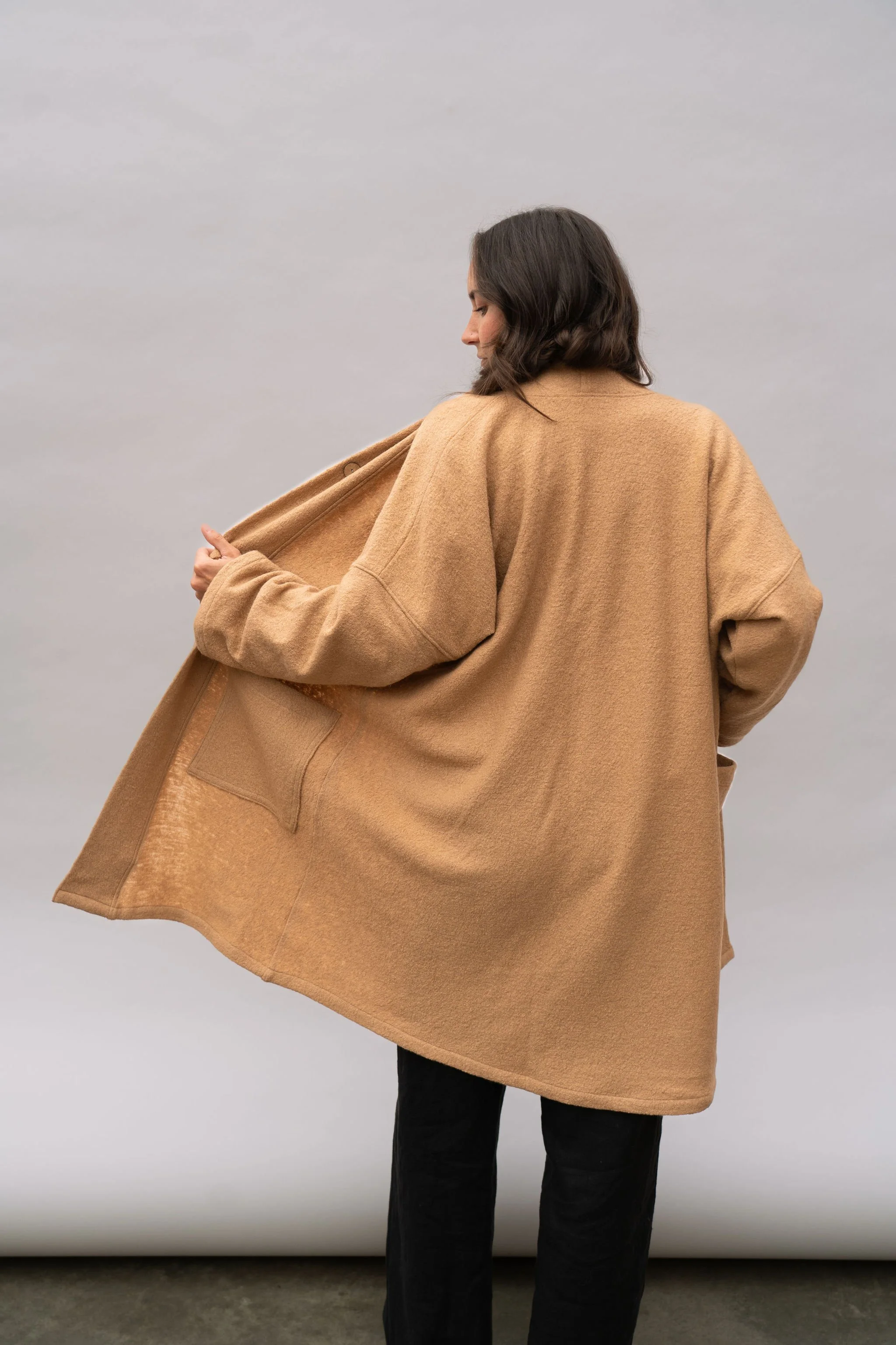 Back view of a woman wearing a wool coat in Camel looking left while holding up the side of the coat