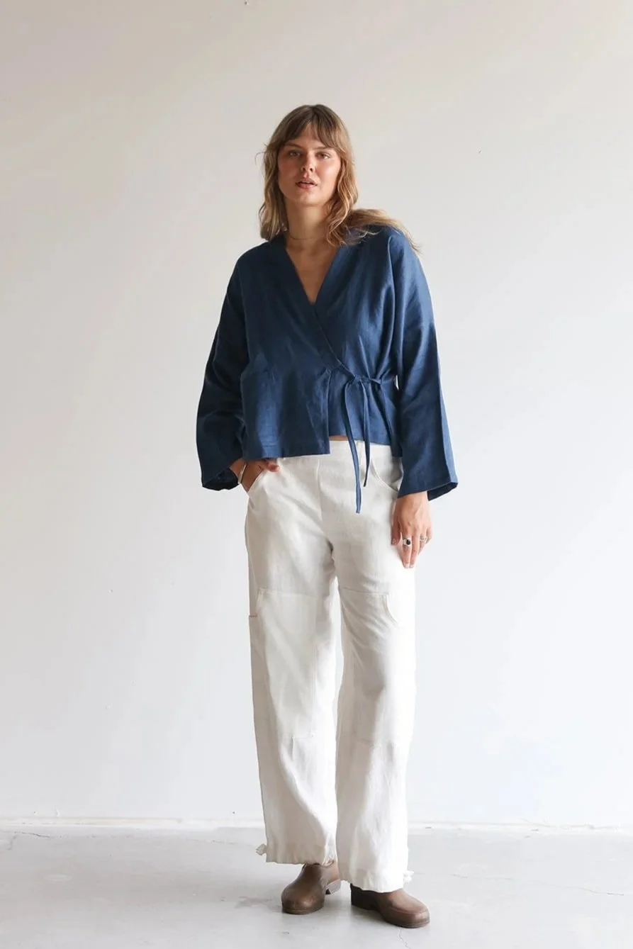 Front view of a woman wearing a linen wrap top in blue with one hand in pocket of white linen pants