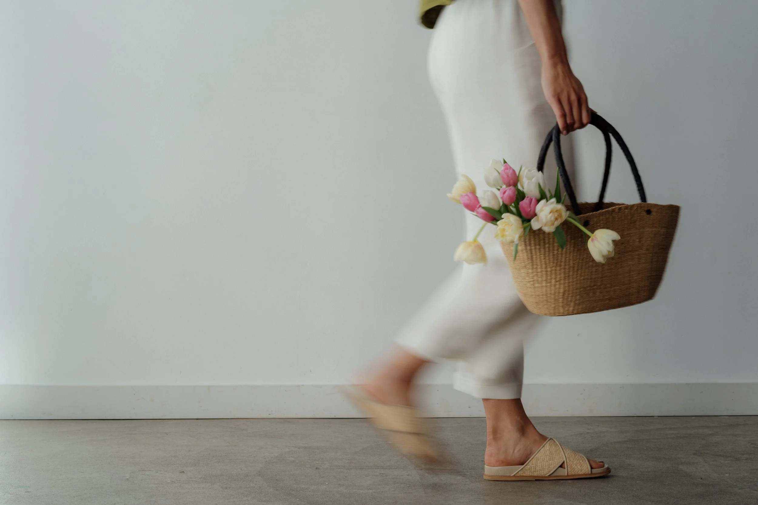 Person carrying a wicker basket with pink and white tulips, walking indoors in cream-colored pants and sandals.