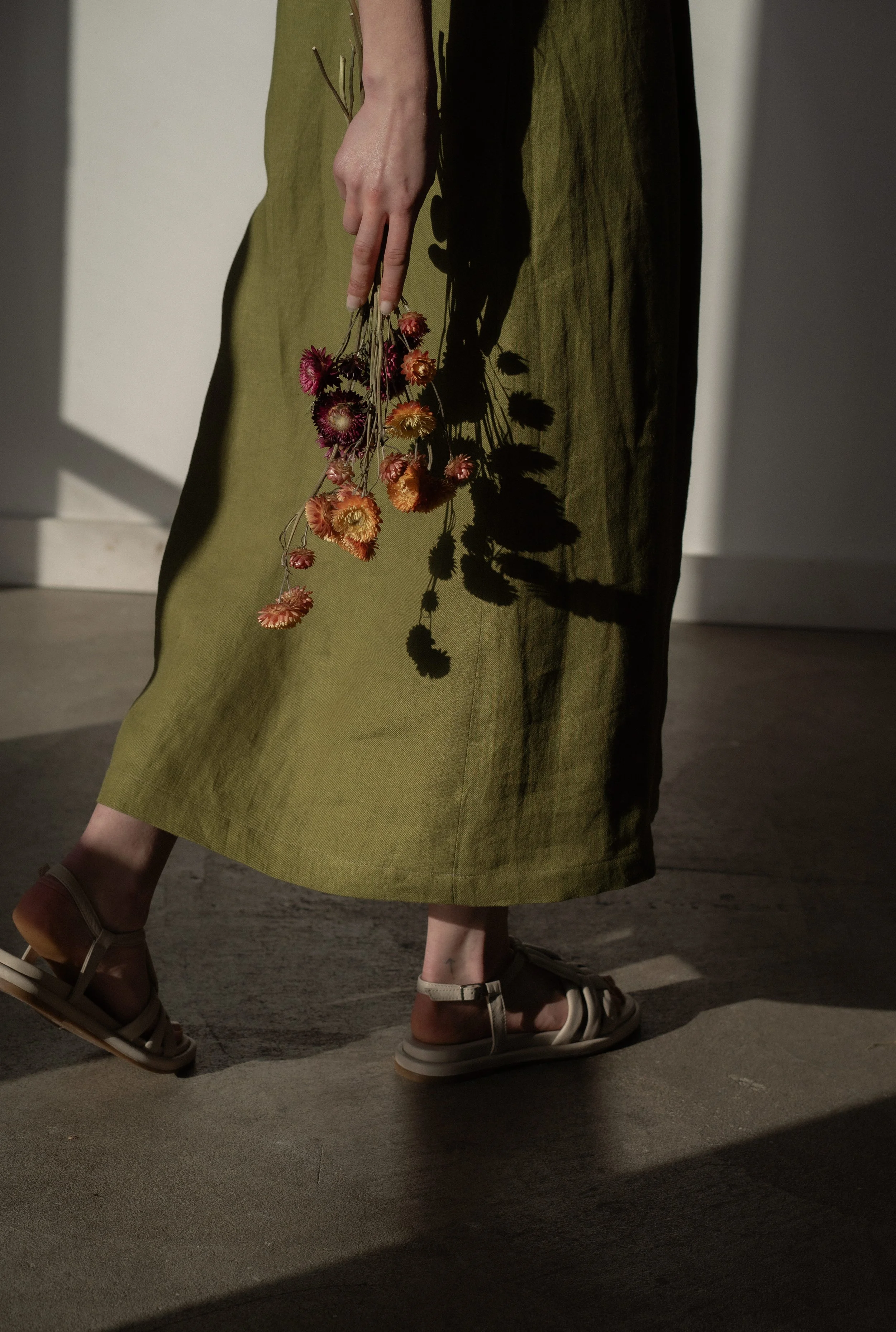 Person wearing an olive green linen skirt, beige sandals, holding a small bunch of dried pink and orange flowers, with the shadow of the flowers cast on the skirt.