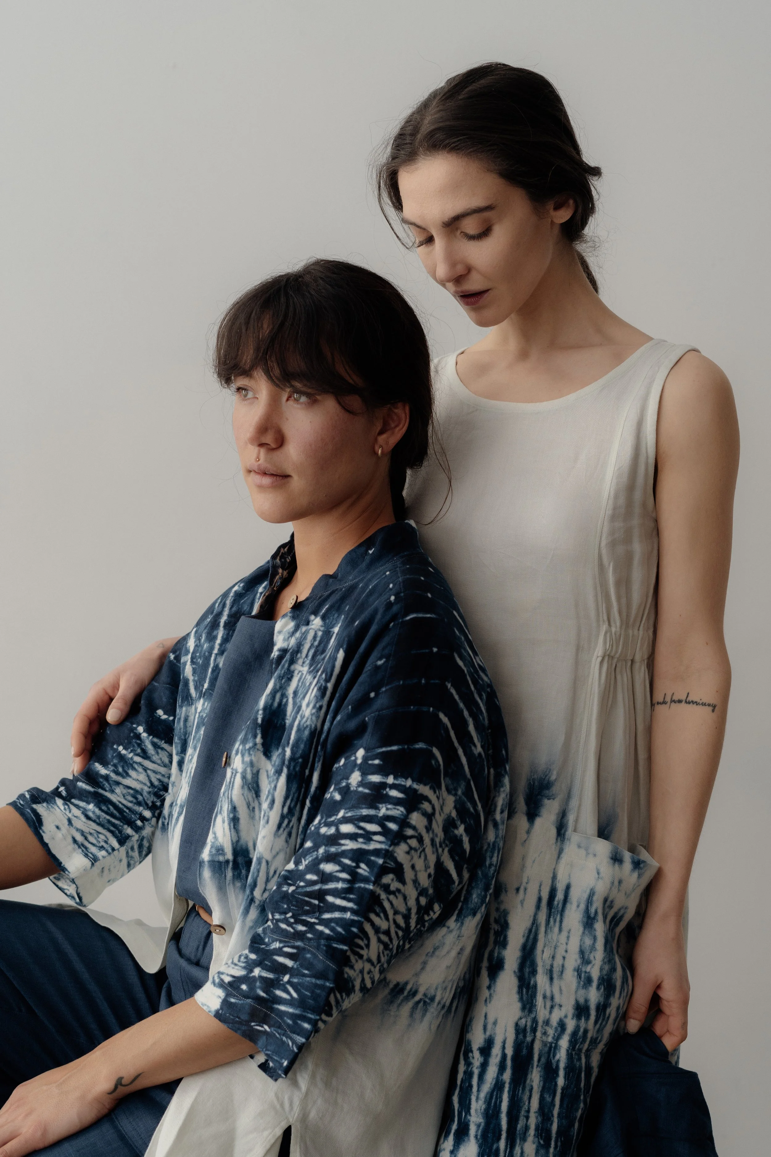 Two women are posed together against a plain background, one sitting and the other standing close behind her. The woman sitting has short dark hair and wears a blue and white patterned robe-like garment. The woman standing has long dark hair, wears a