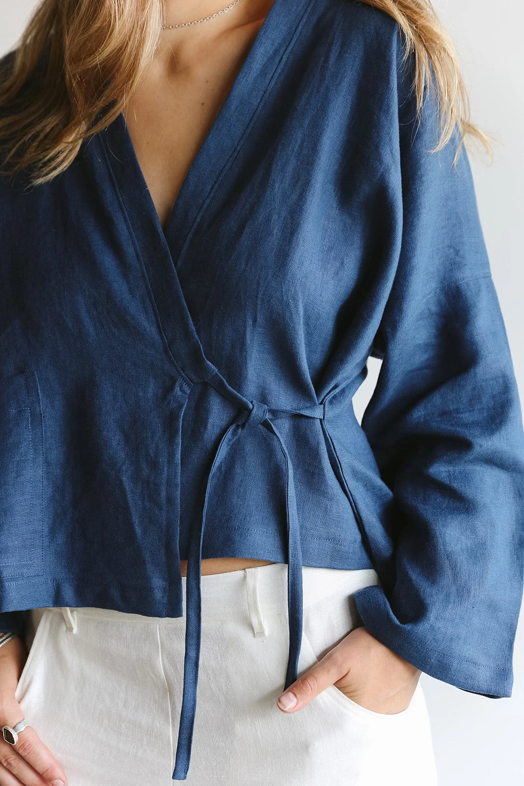 Close up of a woman wearing a linen wrap top in blue with hands in pockets