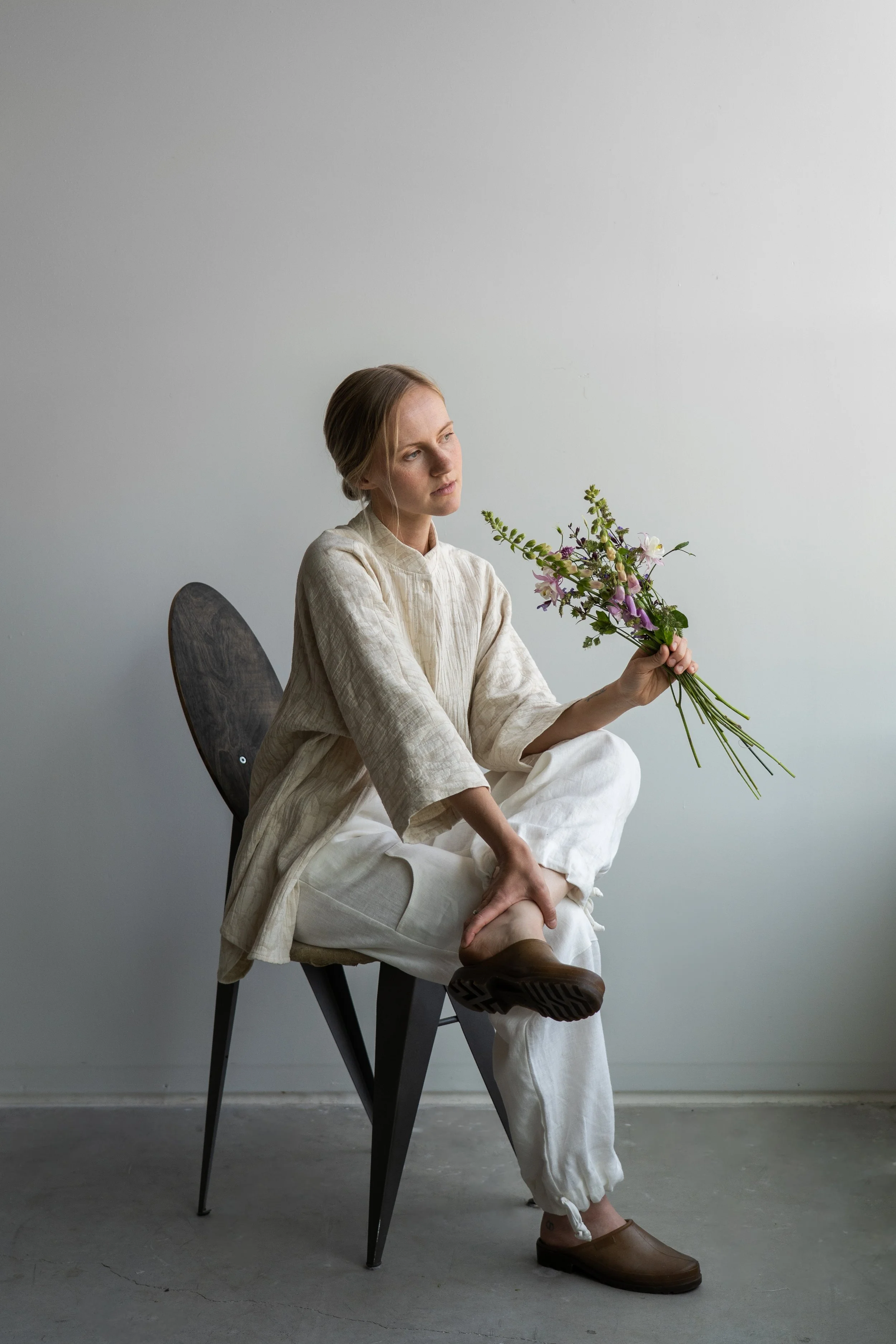 A woman with blonde hair sitting on a black chair in a minimalist room, holding a bouquet of flowers and looking thoughtfully to the side.