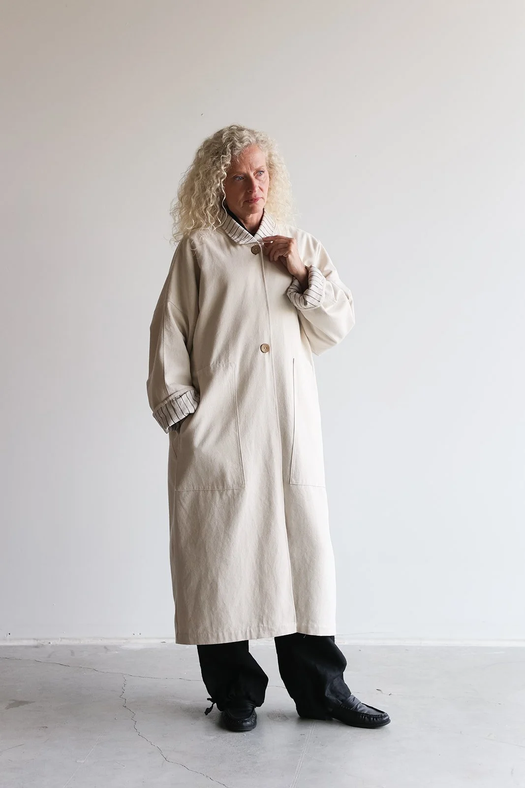 Front view of a woman wearing a canvas linen trench coat in Sand adjusting collar with hand in pocket looking right