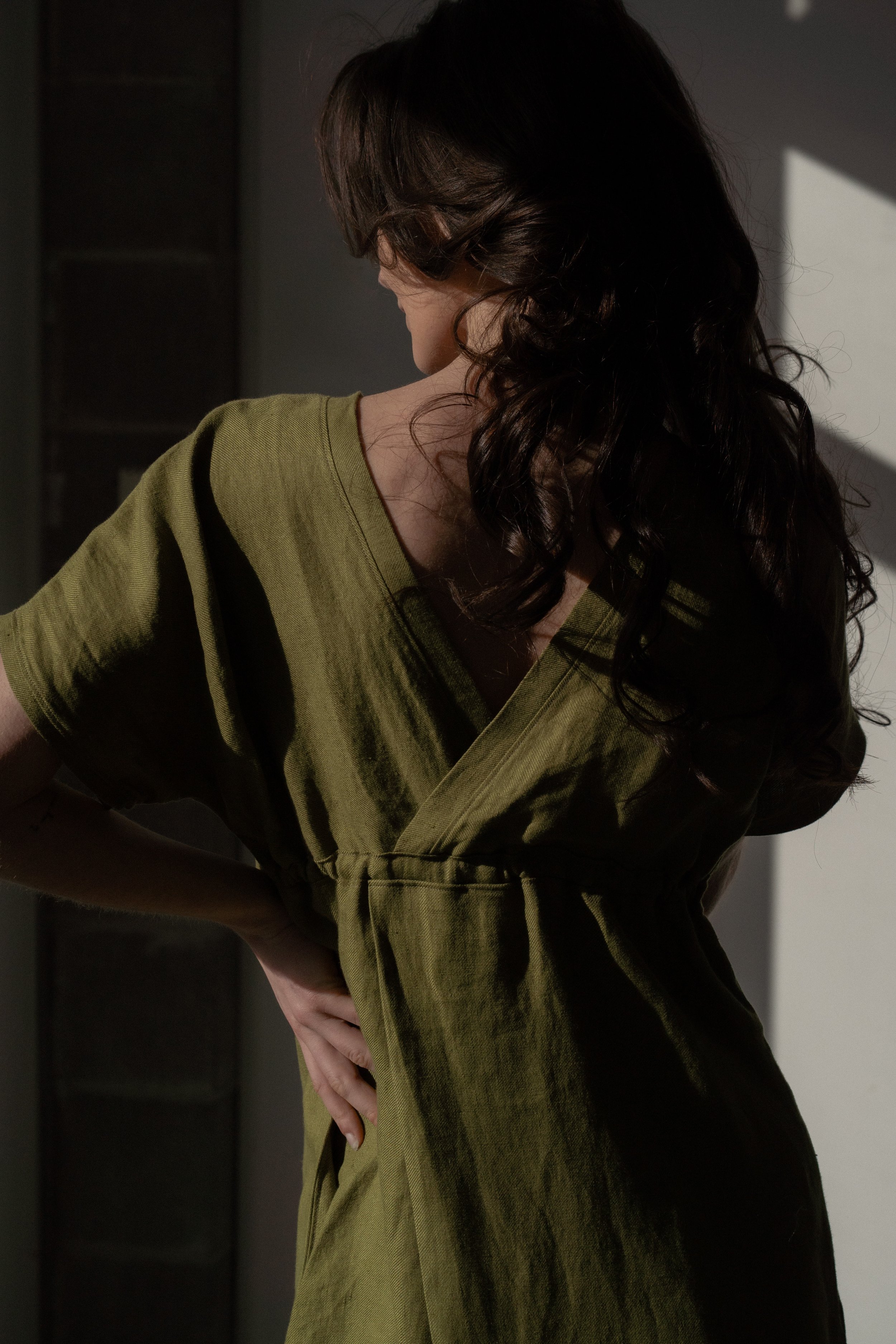 A woman in a green dress standing in shadow with her hand on her back, facing away from the camera.
