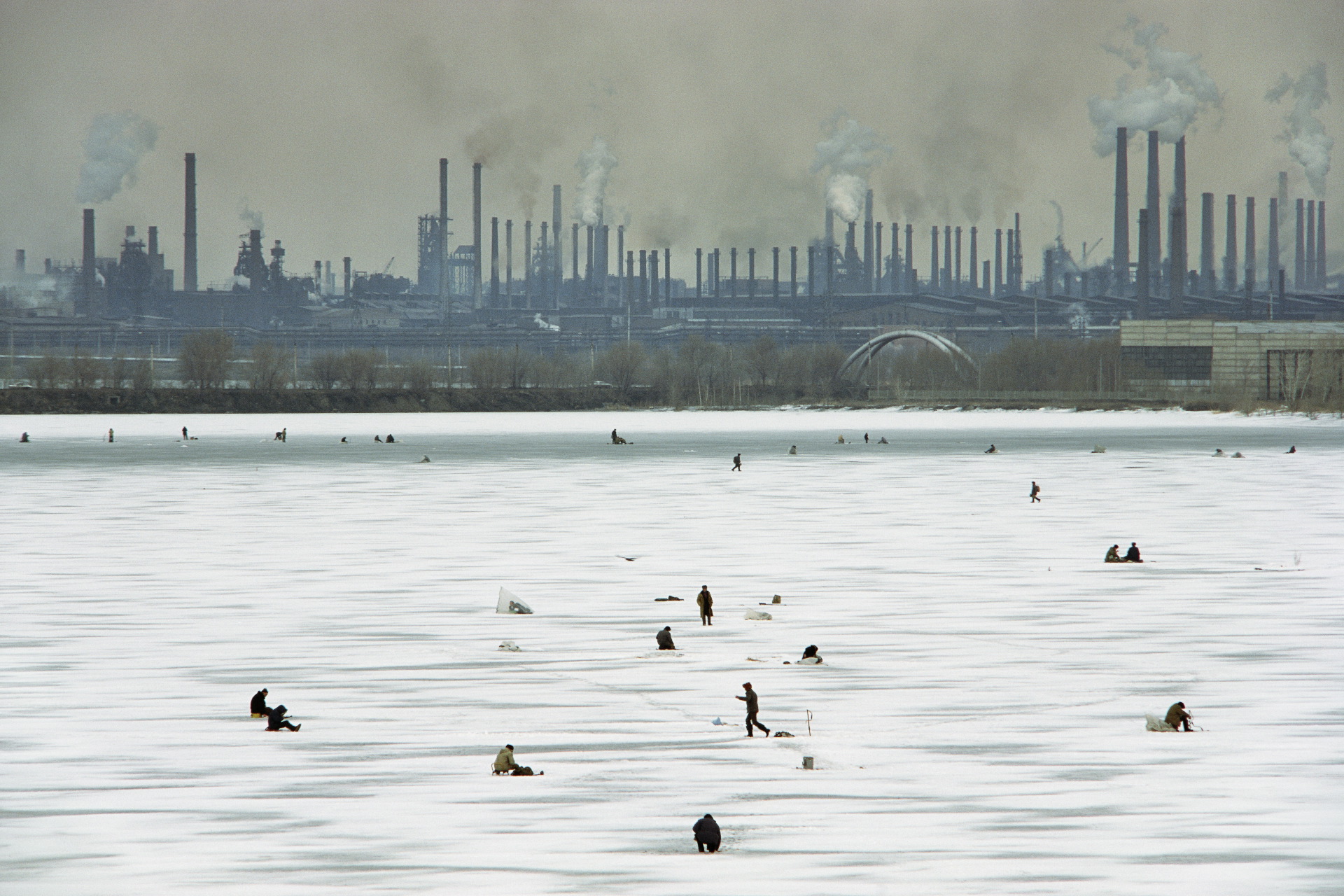  In winter, men drill fishing holes in the ice of the Ural River. Knowing that the river is badly polluted by the Steel Works looming behind them, they often sell their catch to markets rather than consume it themselves.  Magnitogorsk, Russia  