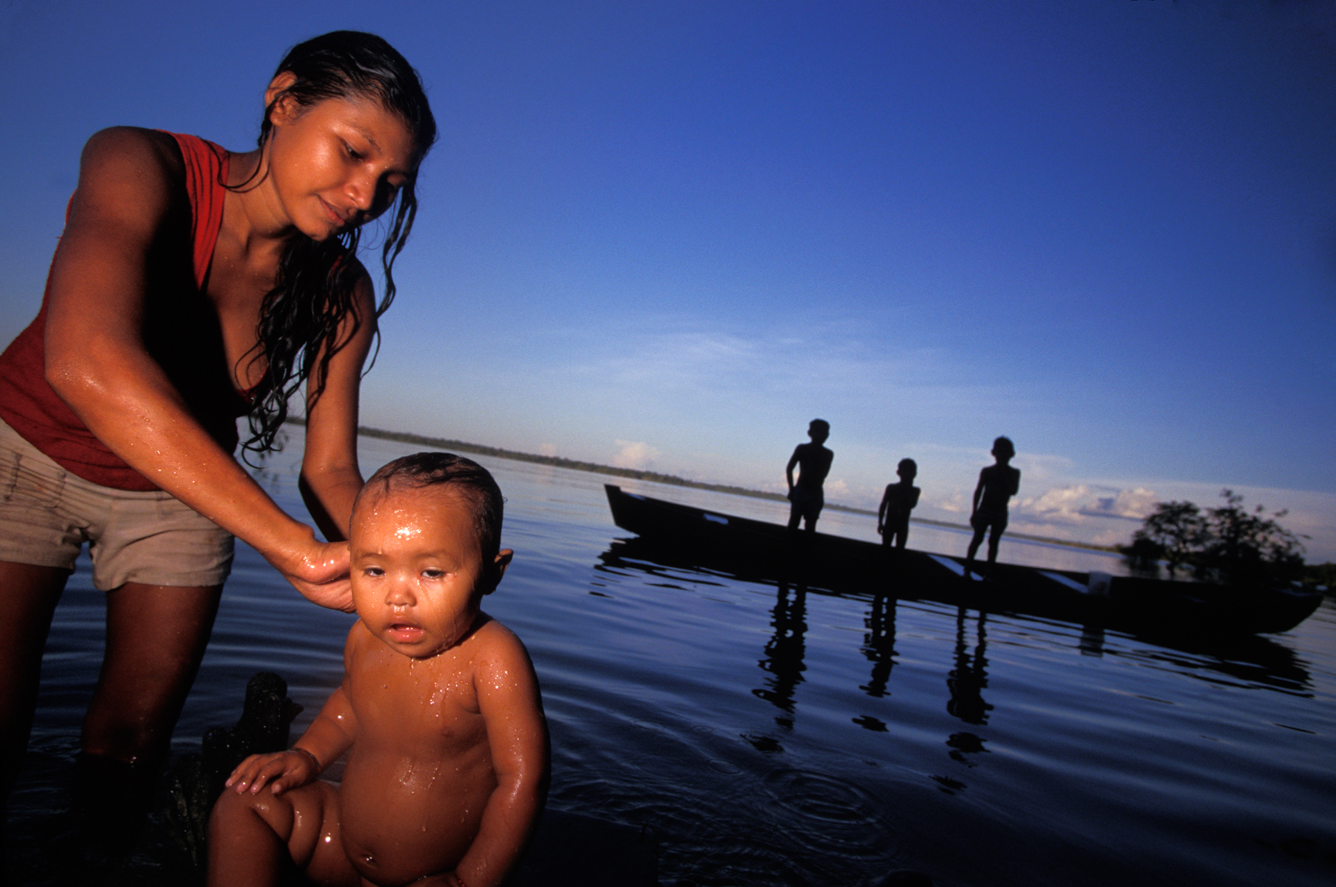  A mother washes her baby in the Rio Canuma while the older kids play in a dugout canoe.  Kwata, Brazil  