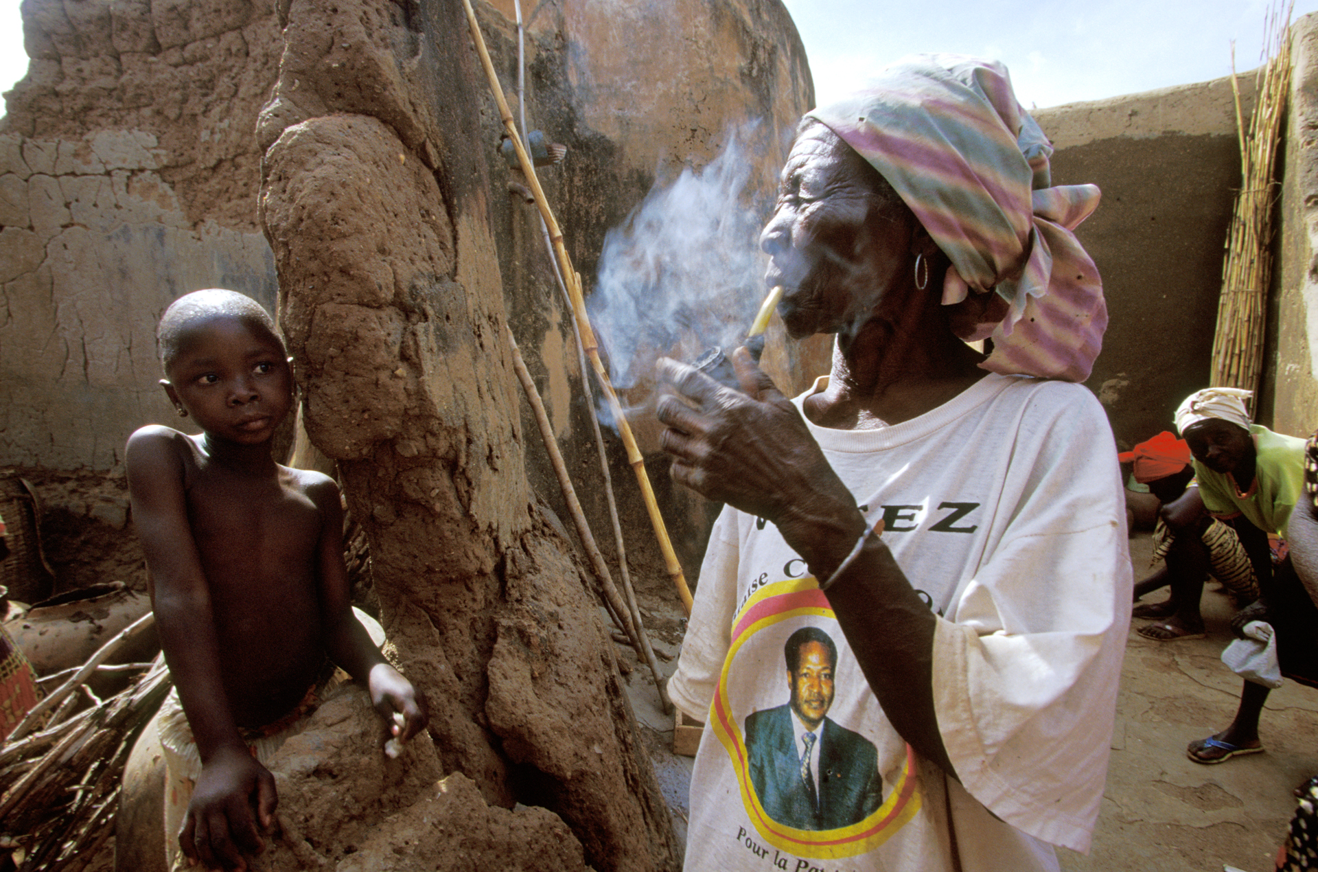  A woman wearing a shirt that identifies her as a supporter of President Compaore takes a break to smoke a pipe during 'House painting day' in the village of Tiebélé.  Tiebélé, Burkina Faso  