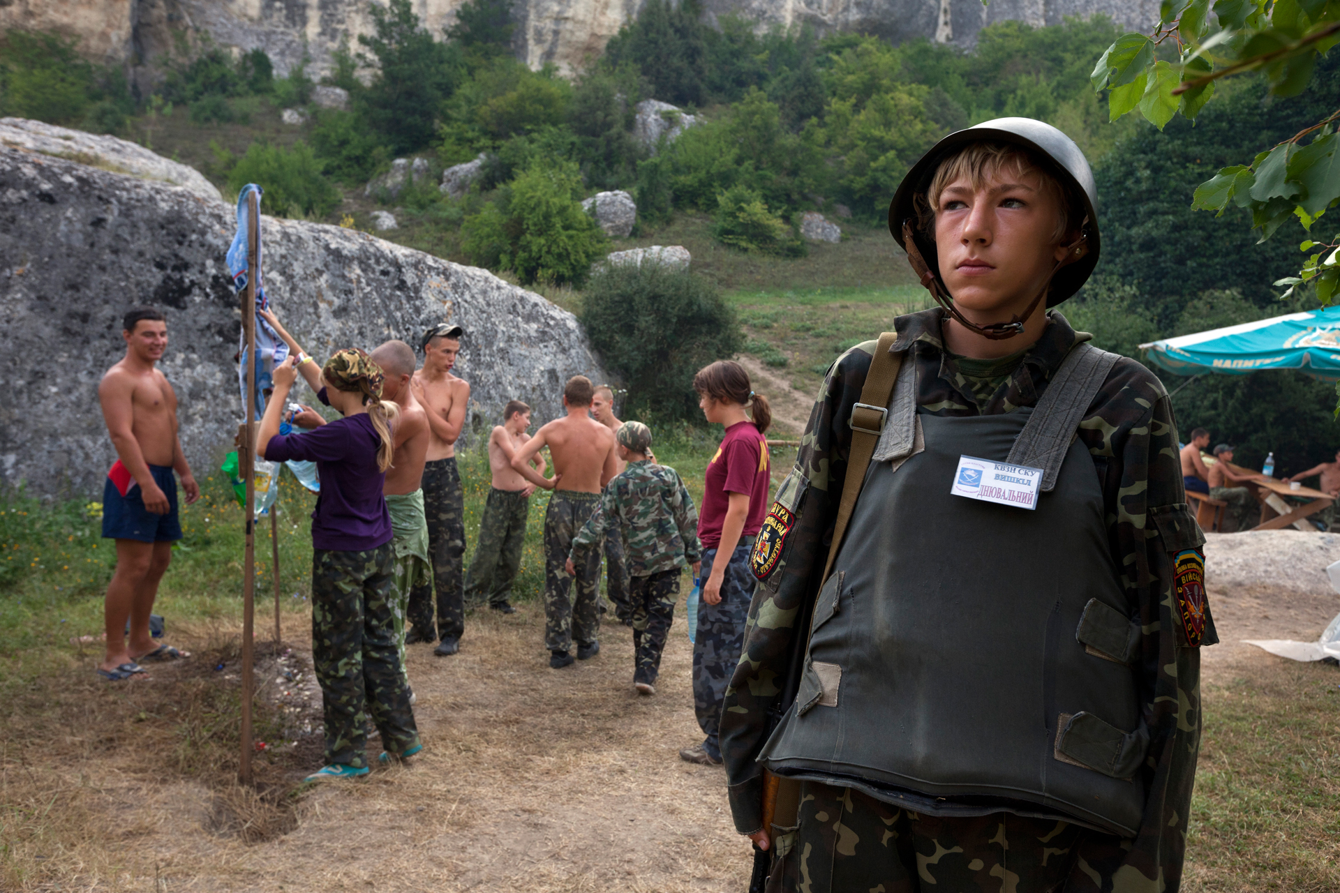  An armed child patrols the entrance to a camp for children aged 8-16, while his peers take care of their morning hygiene.  Eski-Kermen Region, Crimea  