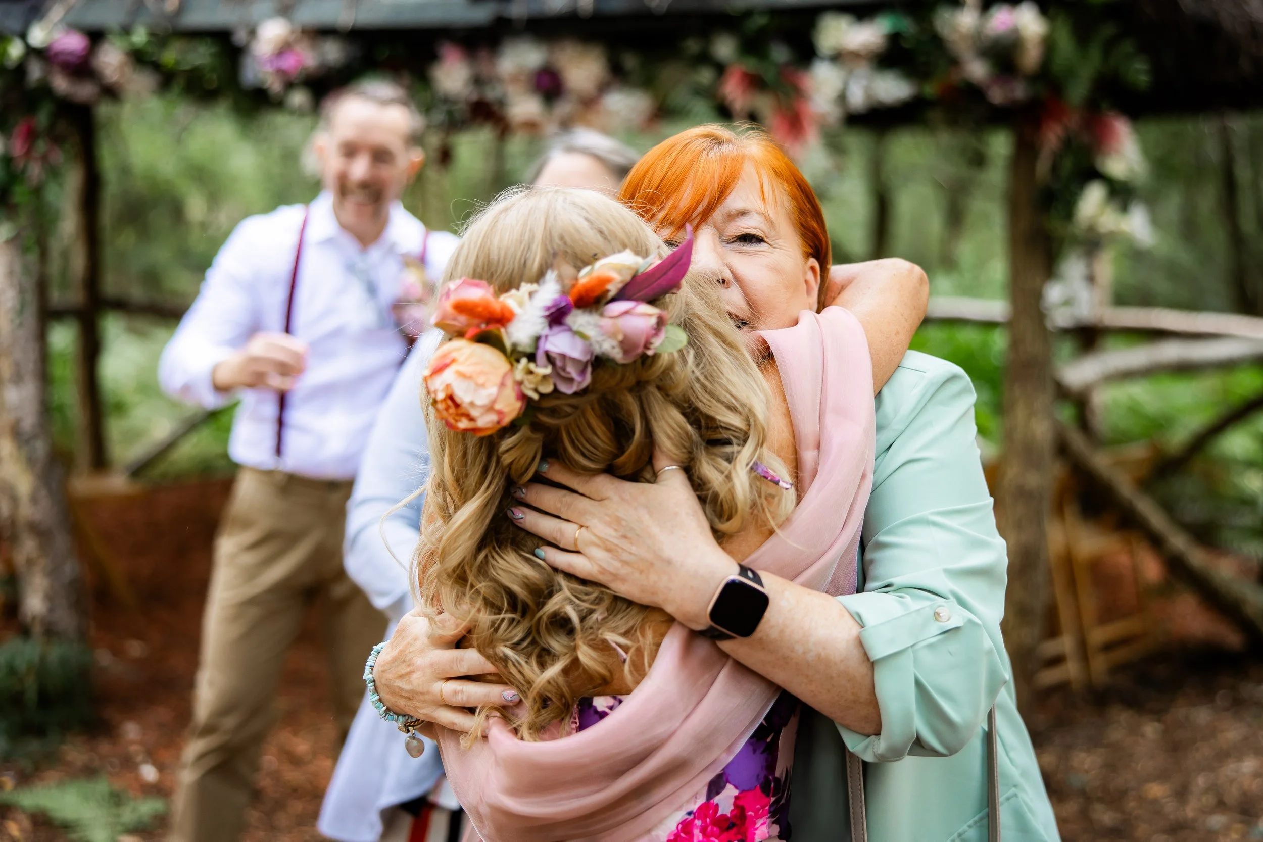 Two women hugging outdoors, one with red hair and one with blonde hair decorated with a floral crown, with a man in the background smiling.
