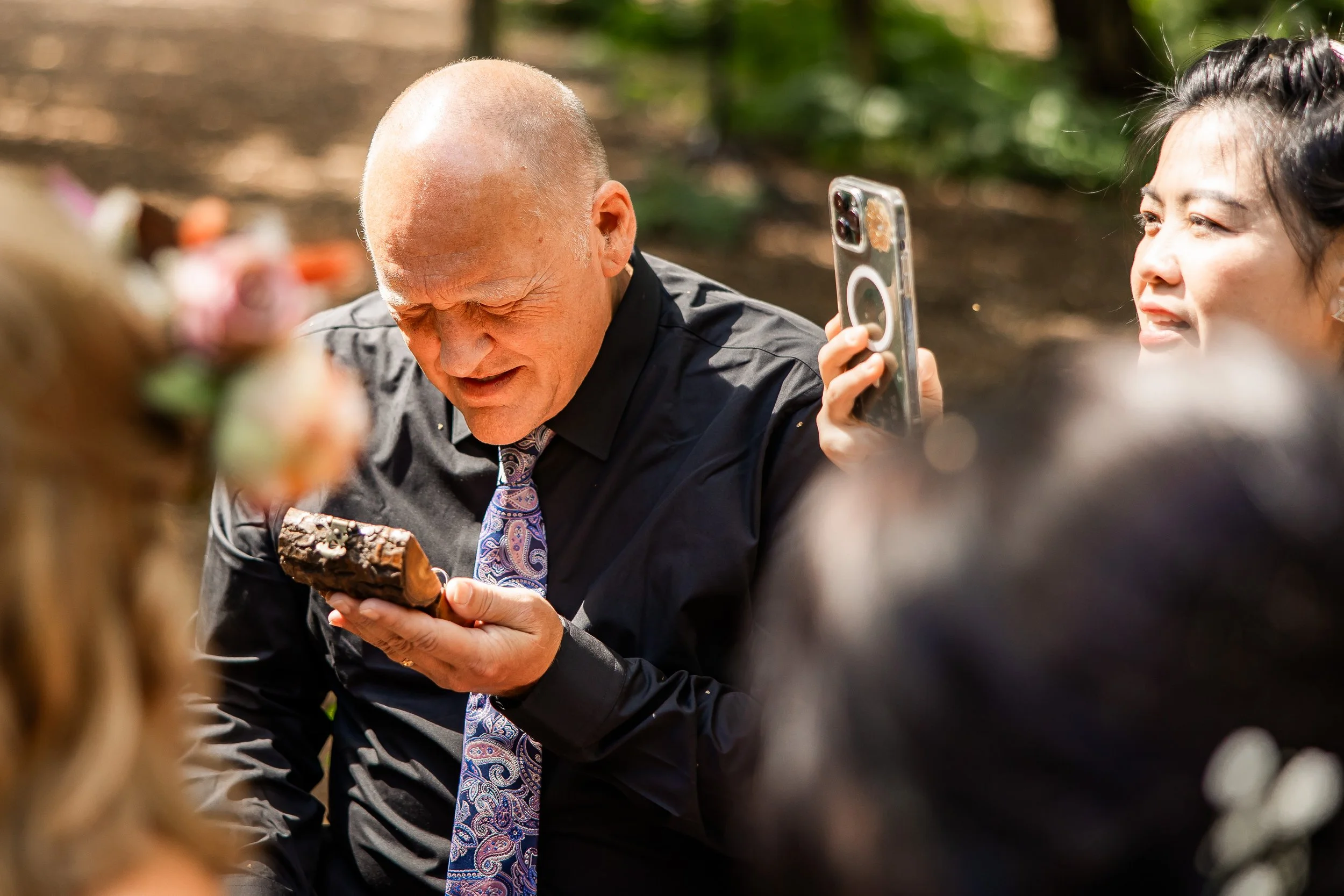 An elderly man is holding a box and appears emotional, surrounded by a group of people. A woman nearby is taking a photo with her phone.