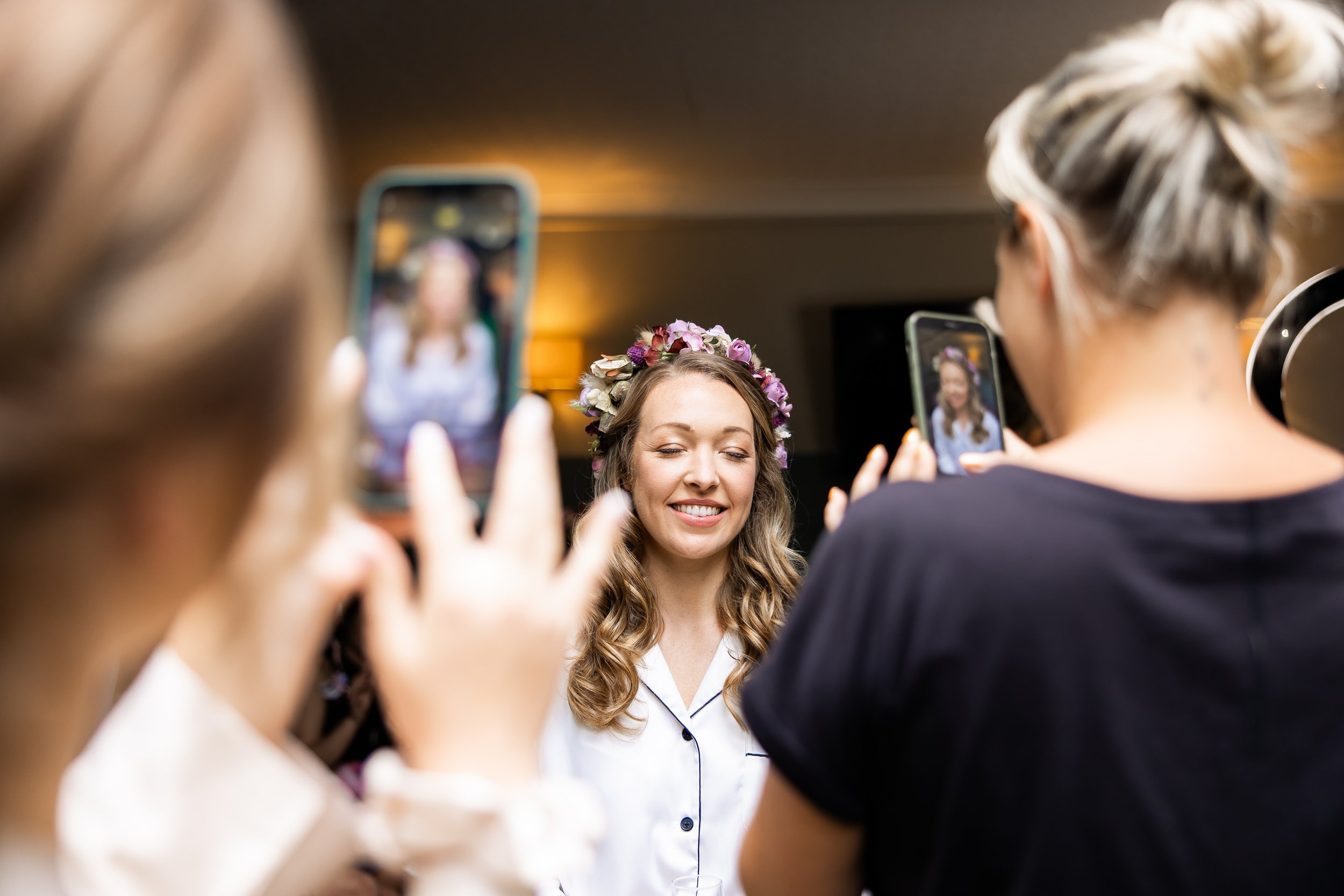 A bride wearing a floral crown smiles with closed eyes as she takes photos with two women using smartphones at her wedding.