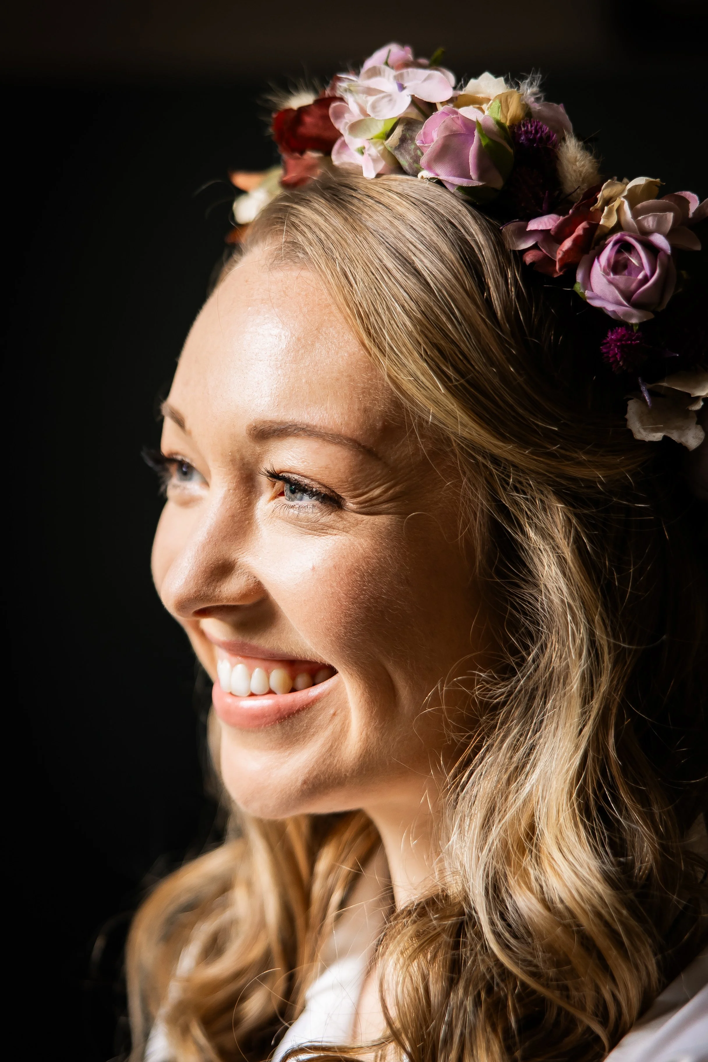 Close-up of a smiling woman with long wavy blonde hair, wearing a floral crown with pink, red, and purple flowers against a black background.