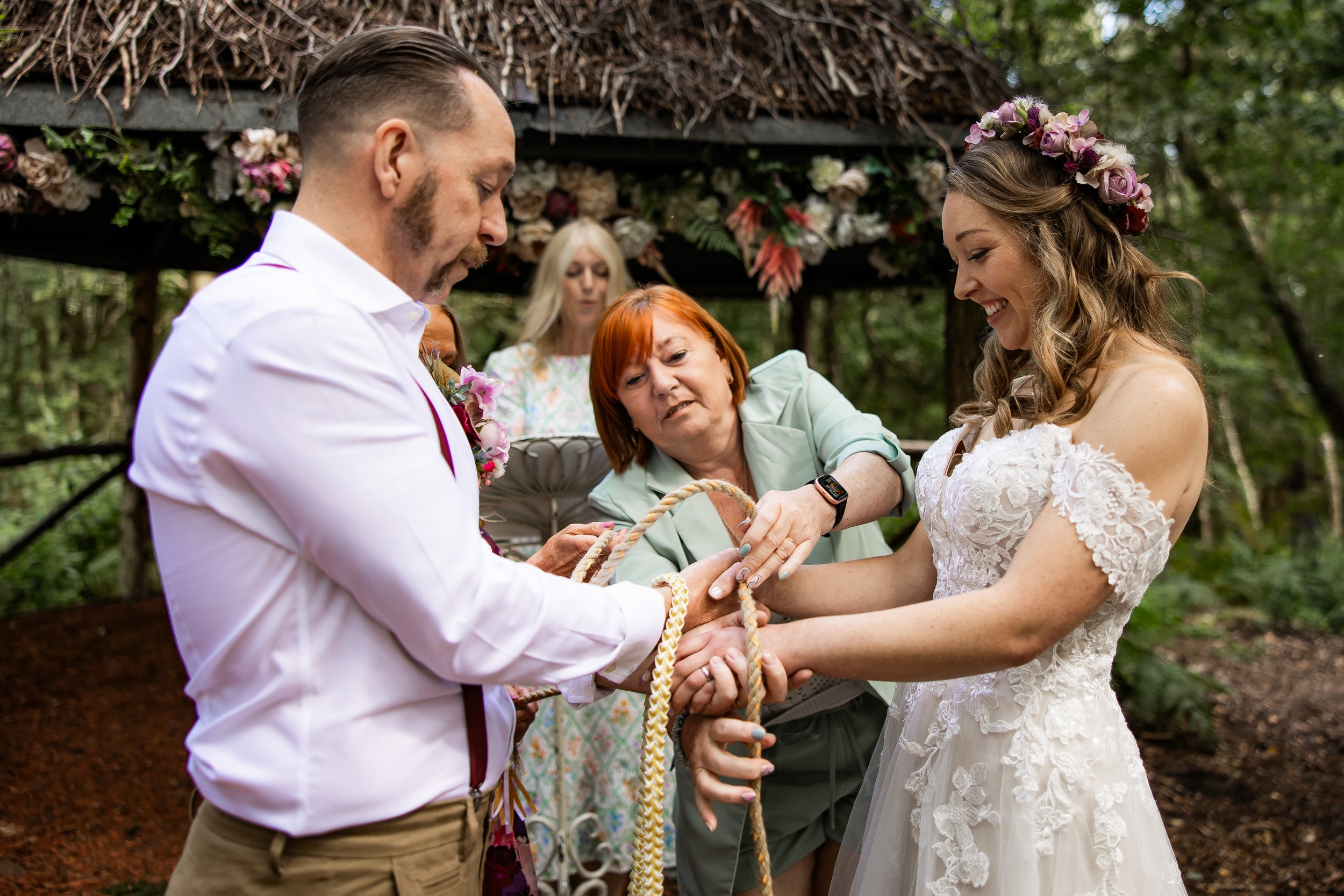 A couple handfasting during an outdoor wedding ceremony, exchanging rings, with a woman officiating and a woman in the background holding a book, under a rustic wood and floral canopy in a wooded area.