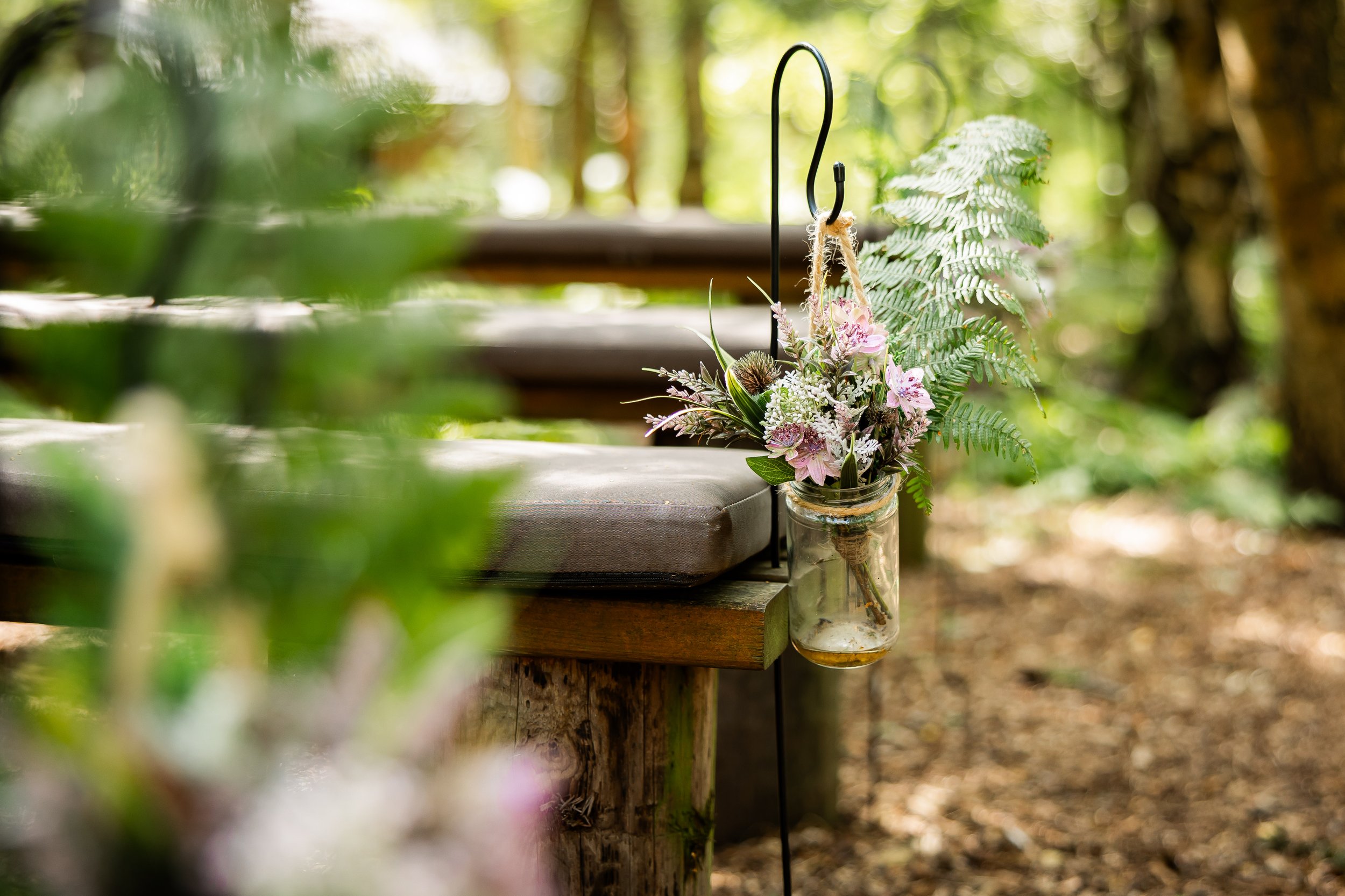 A glass jar with a bouquet of flowers hangs from a hook attached to a wooden bench in a forest setting, with blurred green foliage in the foreground and trees in the background.