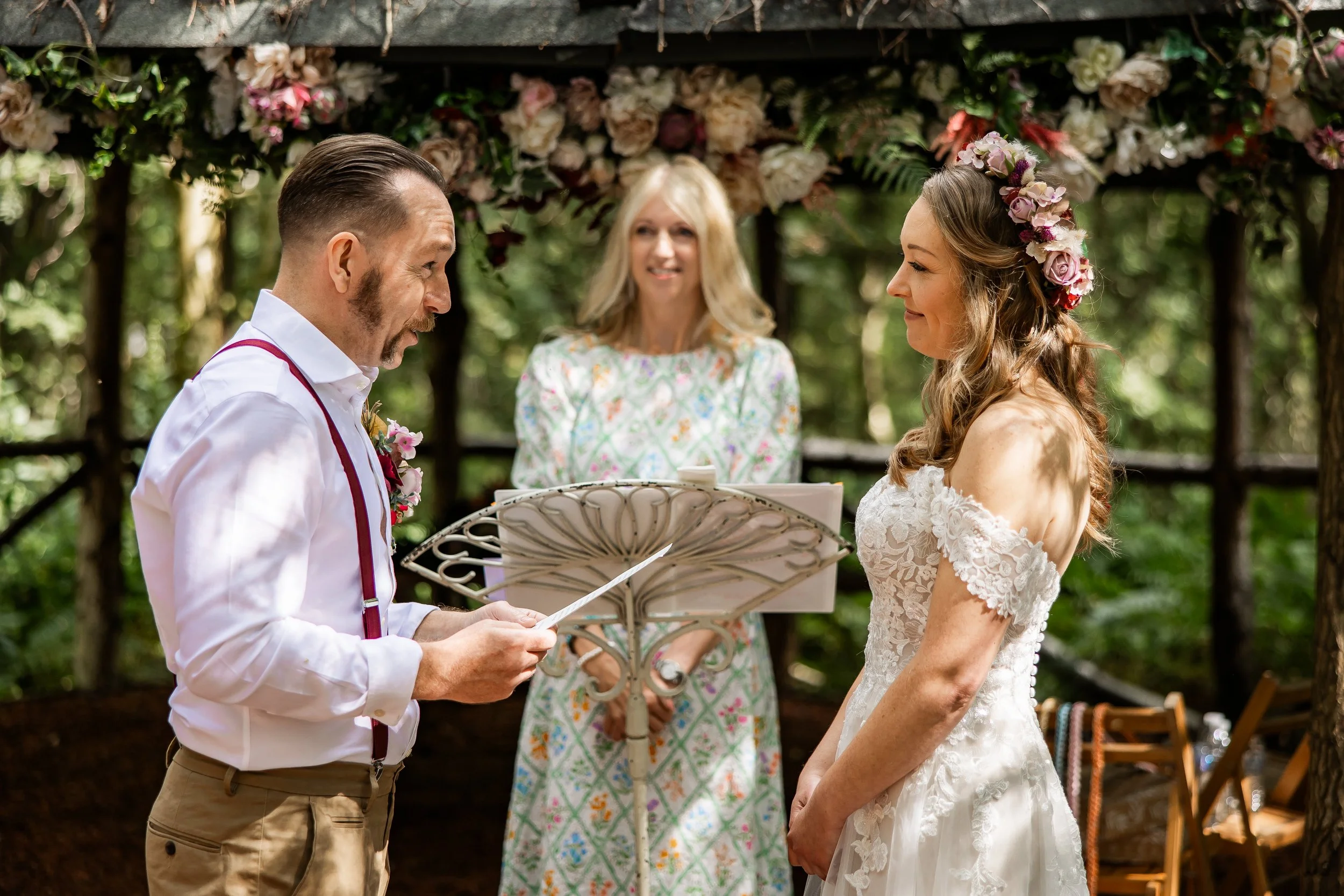 A couple getting married outdoors, exchanging vows, with a officiant standing behind them in front of a floral arch, surrounded by greenery.