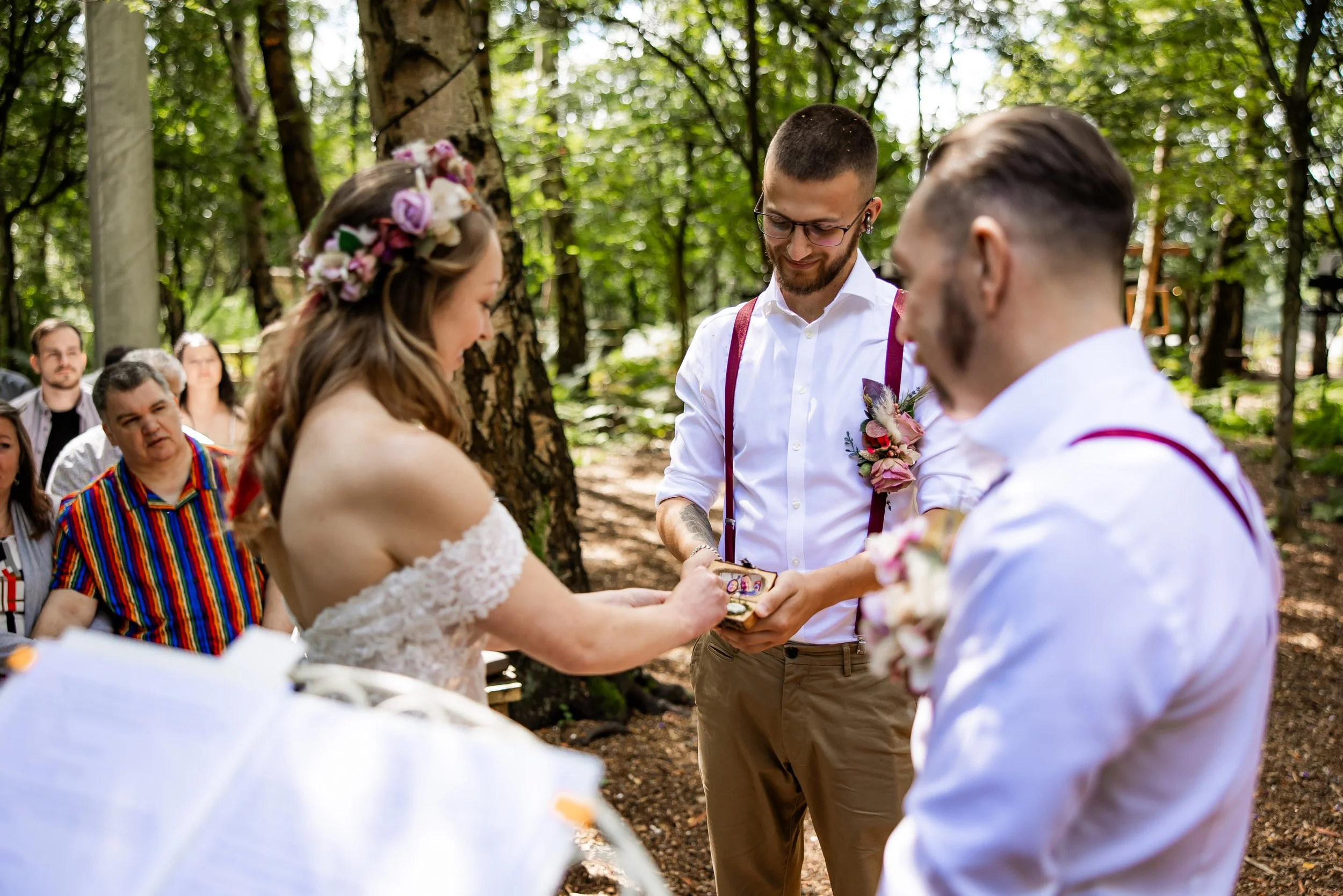 A couple gets married outdoors in a forest, exchanging rings during their wedding ceremony while guests watch.