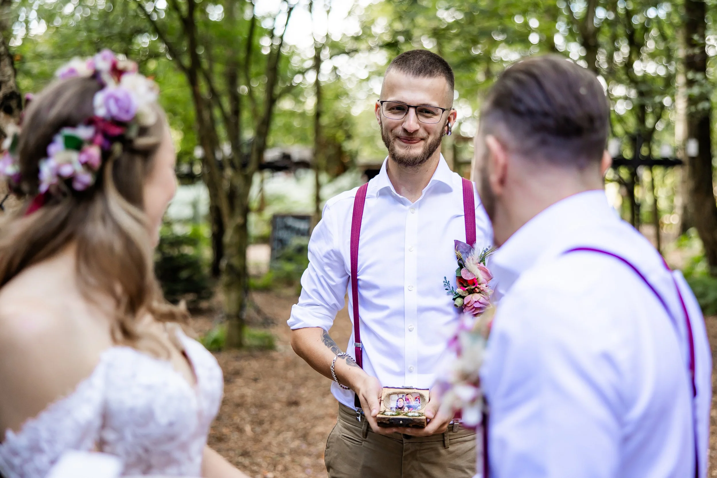 A man holding a box with rings during an outdoor wedding ceremony in a wooded area, with a bride and groom in view.