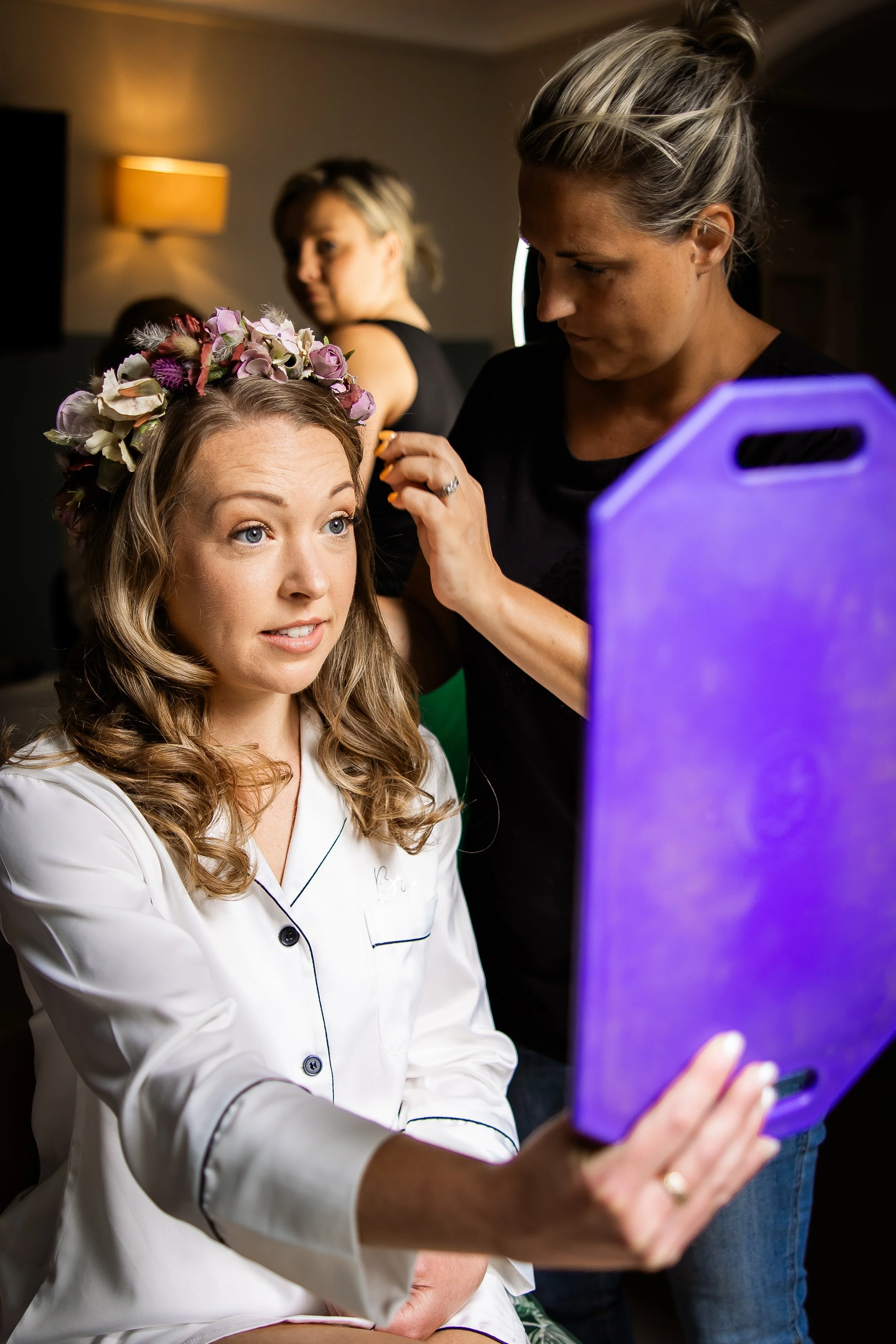 A woman with a floral crown of pink and white flowers sits for makeup or hair styling, with another woman assisting. In the background, another person is visible.