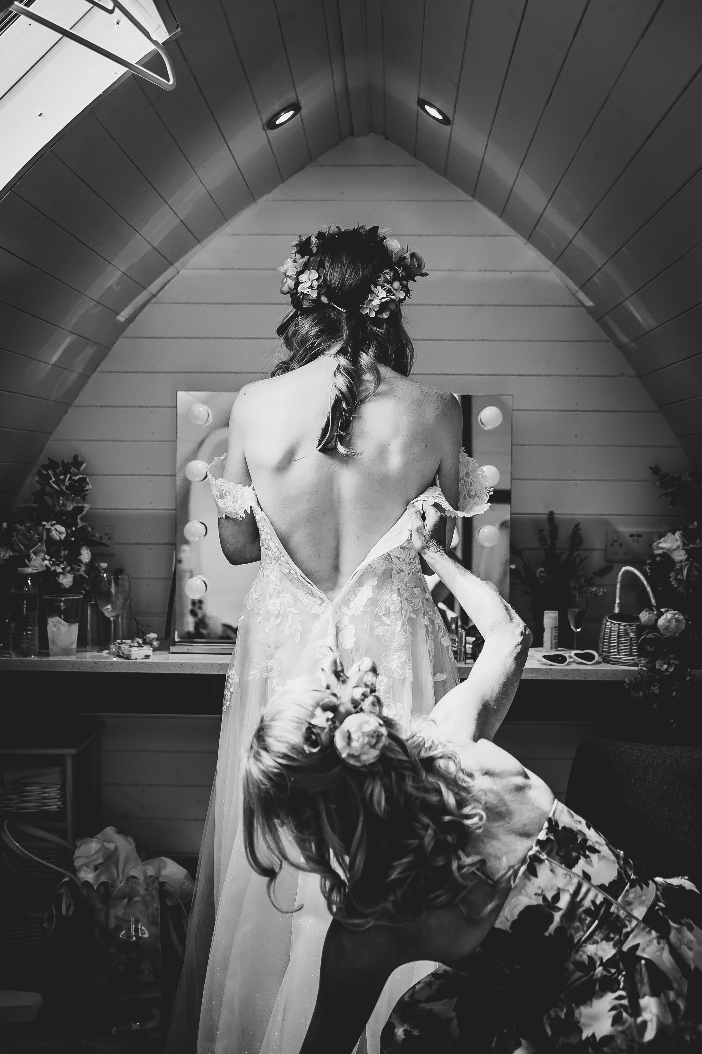 A woman in a wedding dress with her back to the camera, standing in front of a mirror, being helped into her gown by another woman who is kneeling. The woman in the dress wears a flower crown and the scene appears inside a cozy room, possibly a brida