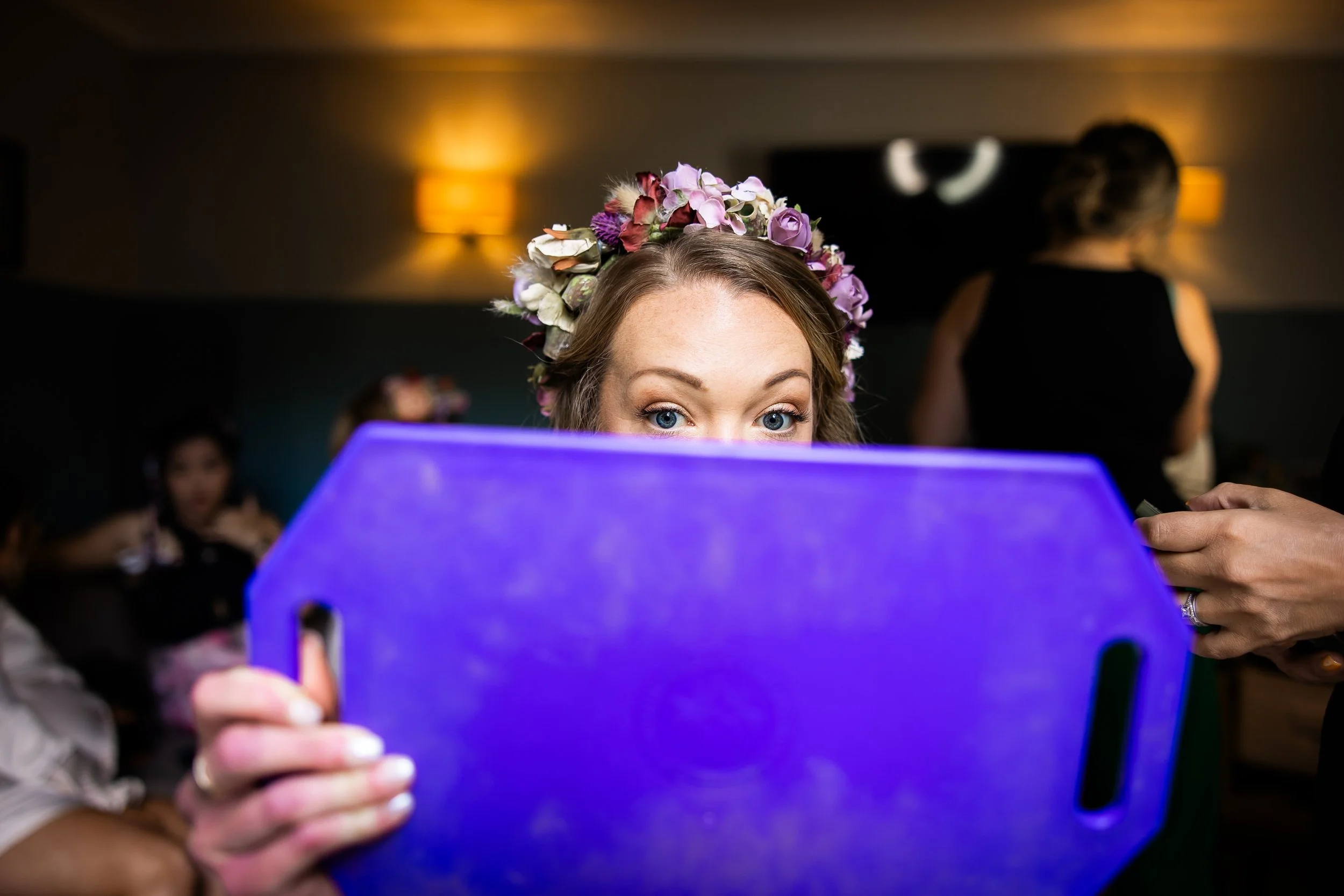 Woman with blue eyes and a flower crown peeking over a glowing blue rectangular object, in a dimly-lit room with other women in the background.