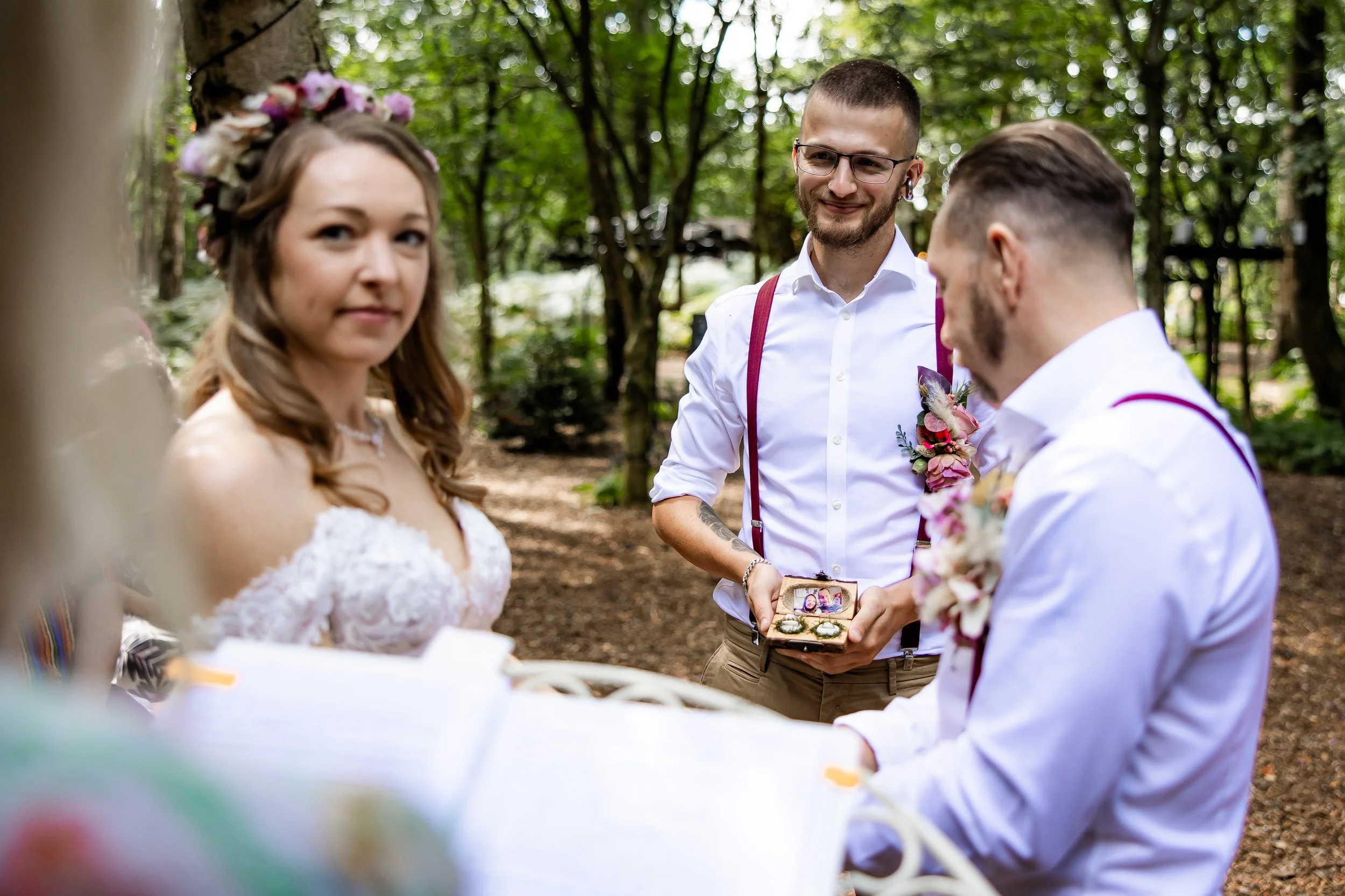A couple getting married outdoors in a forest, with an officiant holding a small open box with rings, and the bride wearing a floral crown and a lace dress.