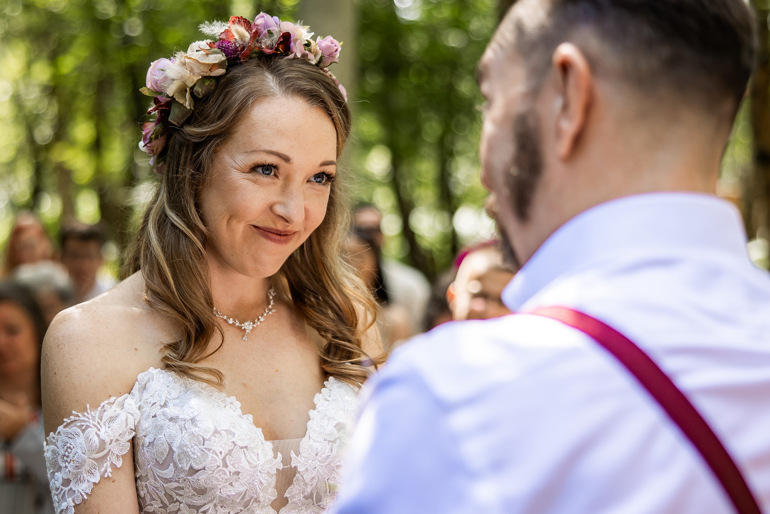 A woman with light brown hair wearing a floral crown and lace wedding dress smiling at a man with dark hair and beard in a white shirt with red suspenders during a wedding ceremony outdoors in a forest.