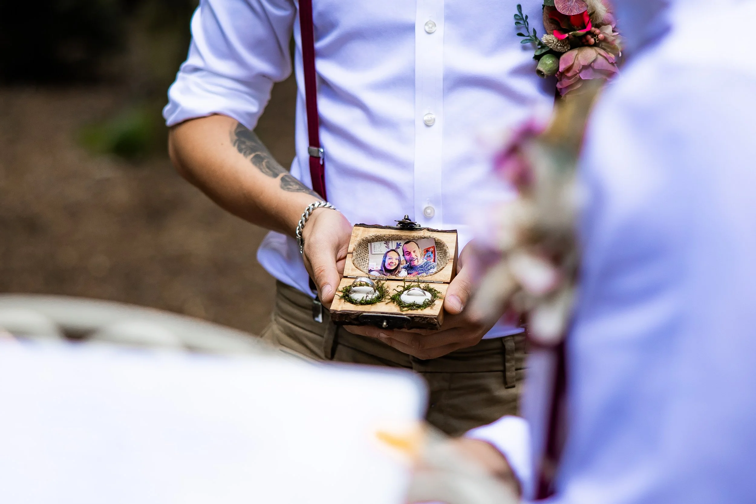 A person holding a small wooden jewelry box with a photo inside and two rings, during a wedding or special ceremony.
