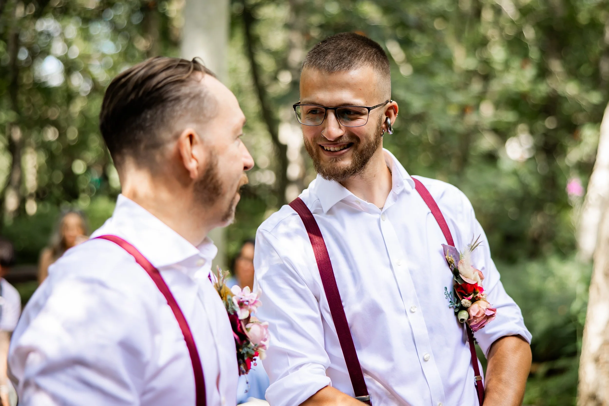 Two men with floral boutonnières and suspenders talking outdoors, surrounded by a wooded area and blurred guests in the background.