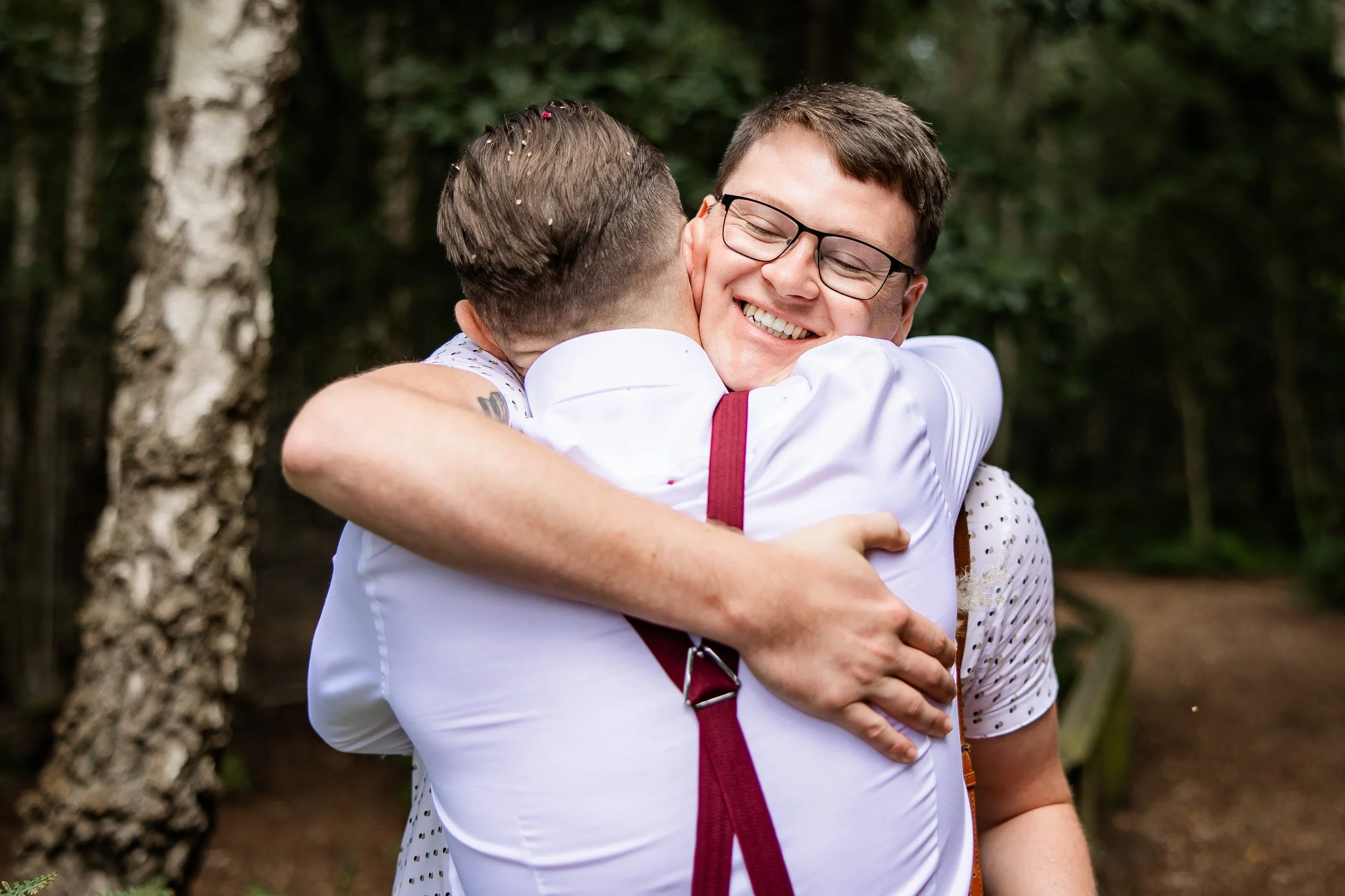 Two men hugging outdoors, one smiling with glasses, in front of a forest background.