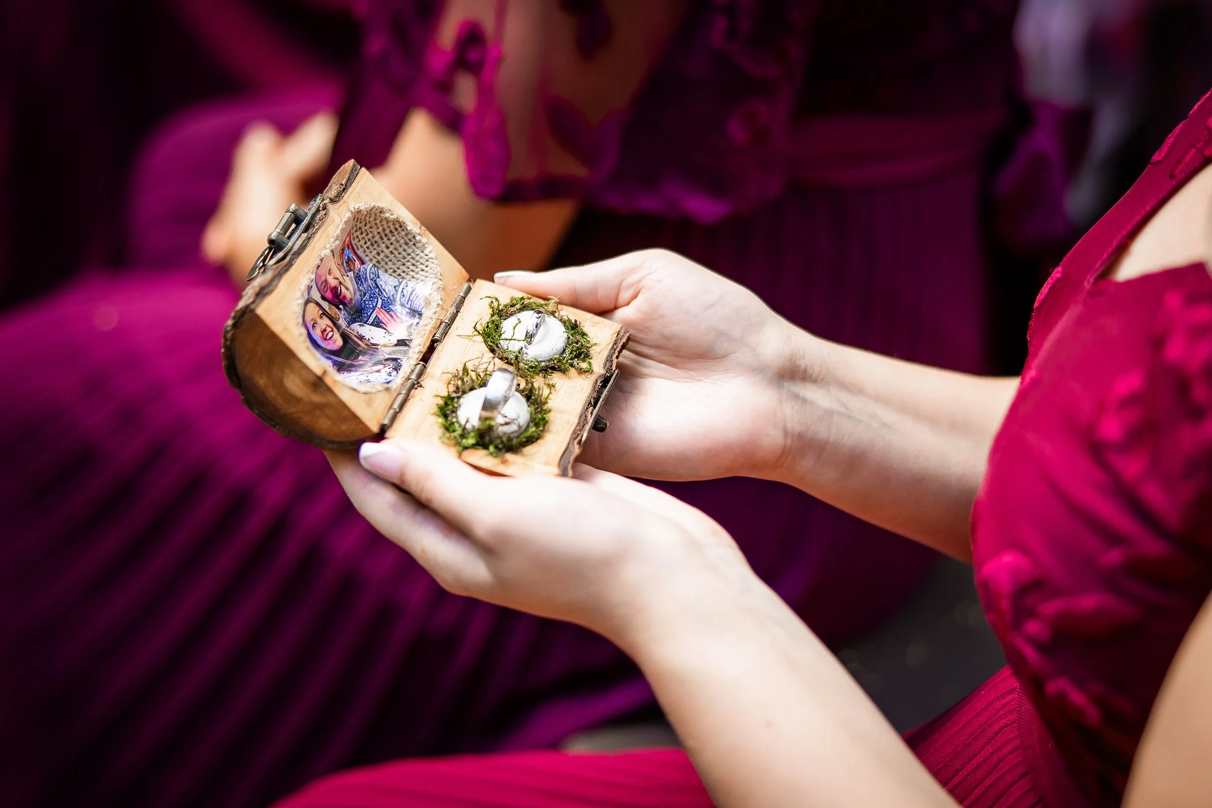 A woman in a red dress holding a small wooden box with two wedding rings and a locket inside, decorated with moss, with a blurred person in purple clothing in the background.