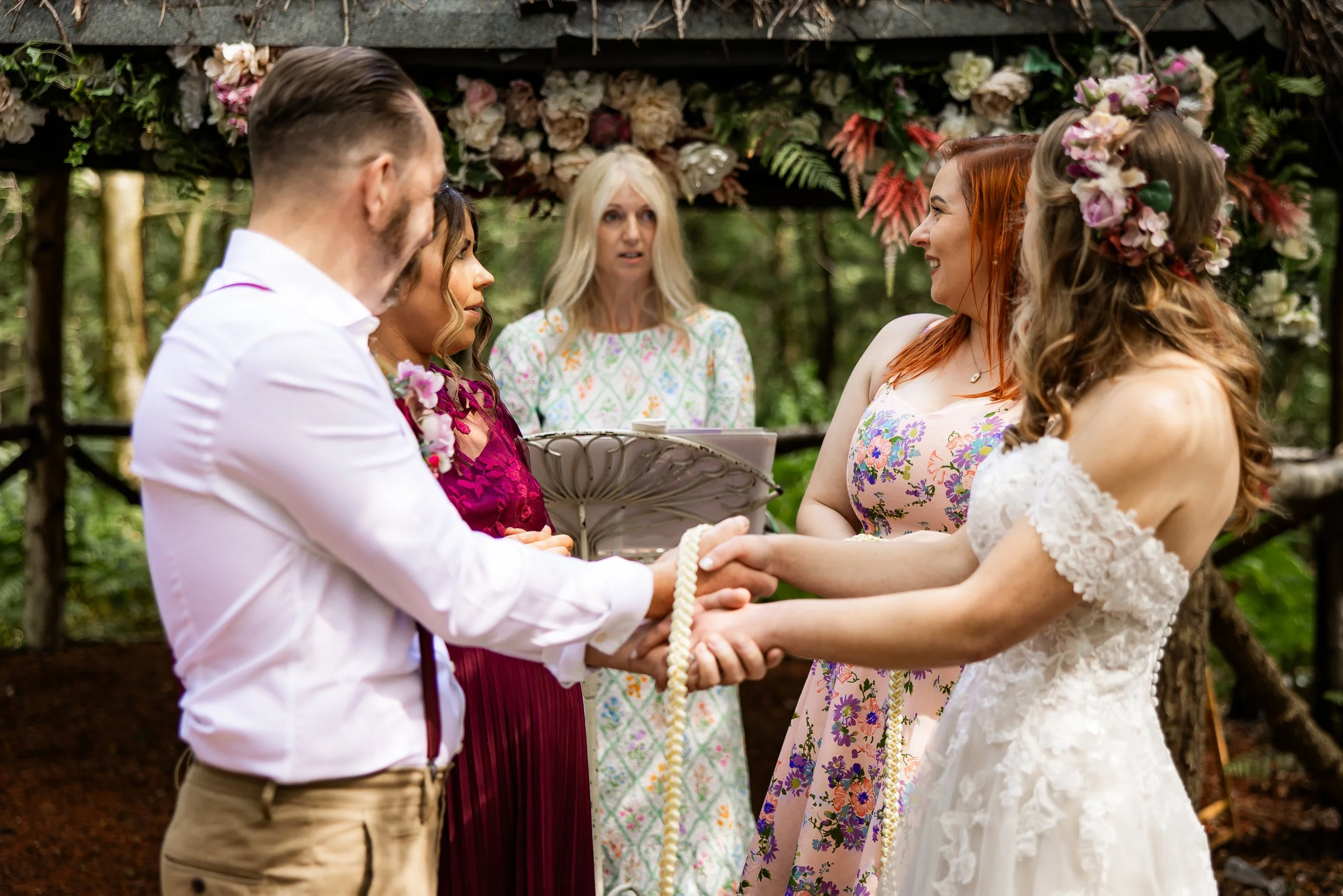 A wedding ceremony with a man and woman holding hands, facing each other, outdoors under a floral arch. An officiant stands behind them with a book as a handfasting ceremony takes place.