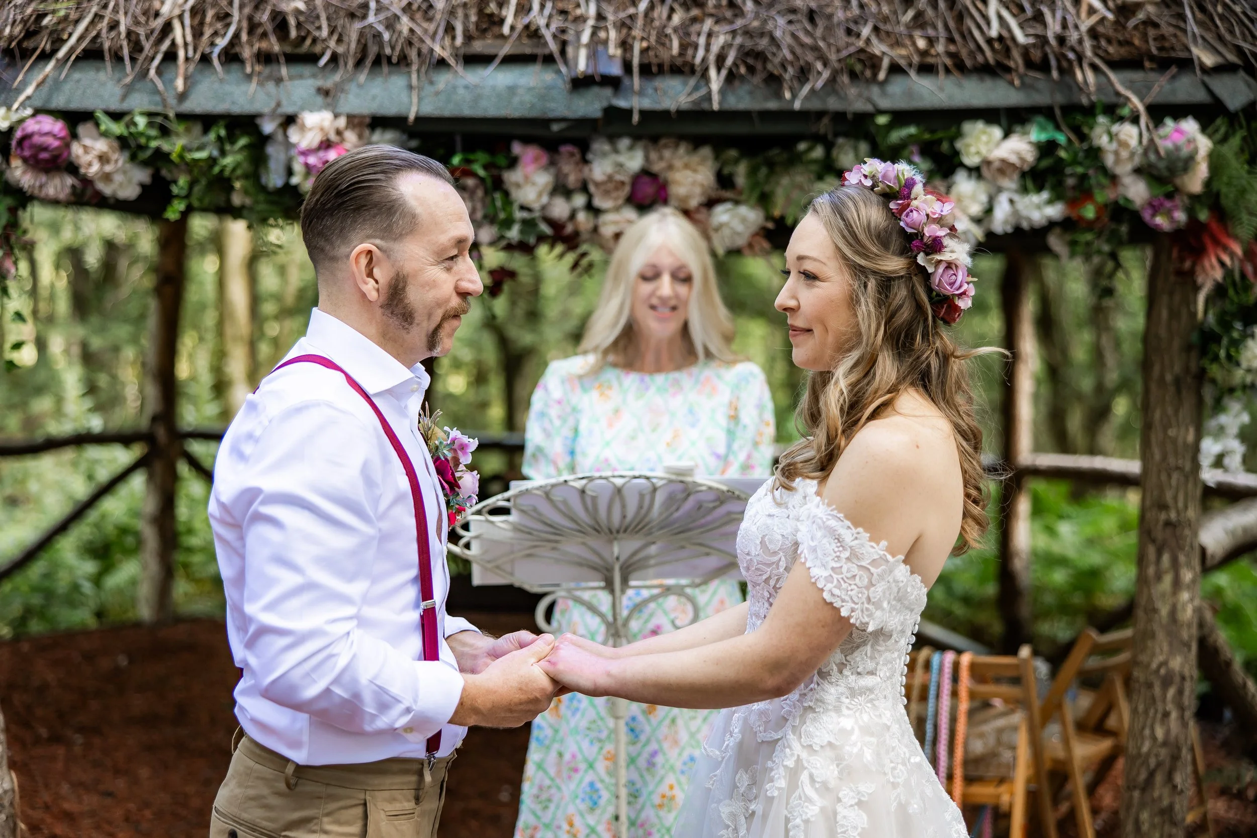 A couple exchanging wedding vows outdoors beneath a floral arch, with a woman officiant standing behind them, surrounded by trees and greenery at Cheshire Woodland Weddings.