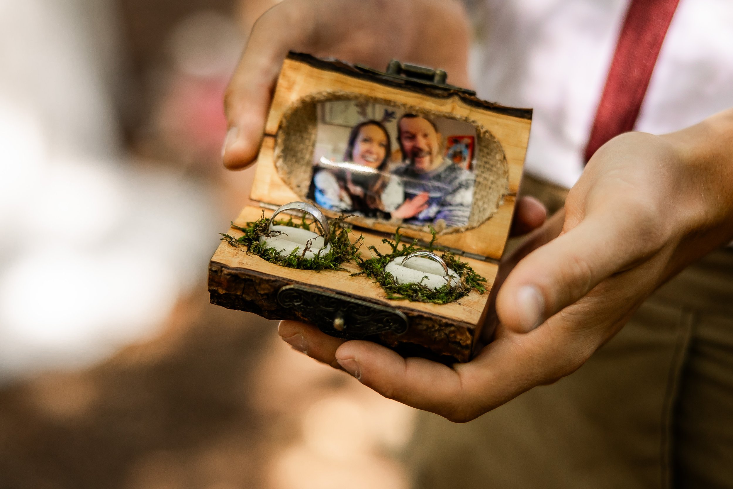 Person holding an open wooden box with a mirror inside, reflecting two smiling people, and two rings with water droplets on moss-lined cushion.