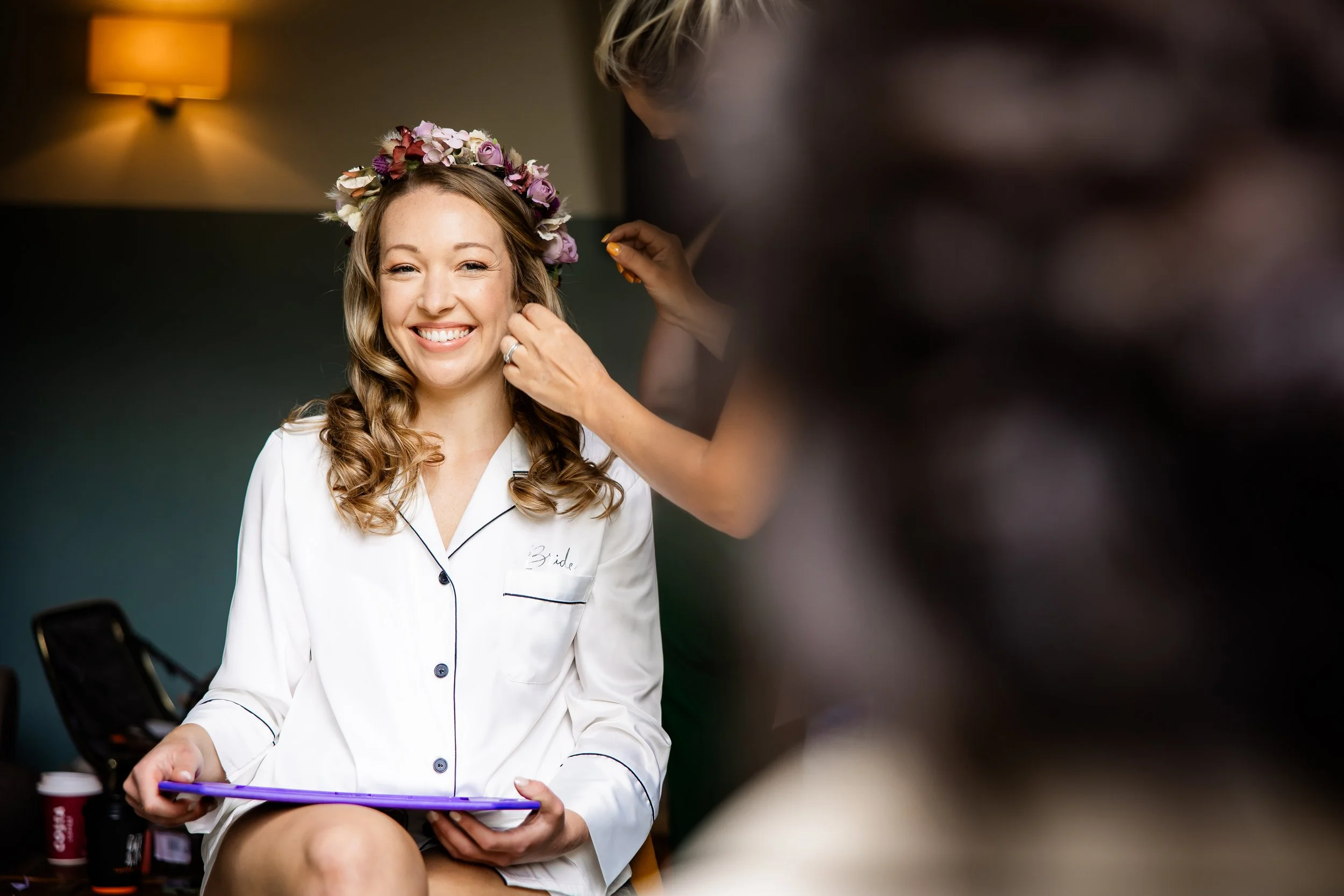 A bride smiling while getting her earrings put on, wearing a floral crown, and sitting in a white pajama top with "Bride" embroidered on it.