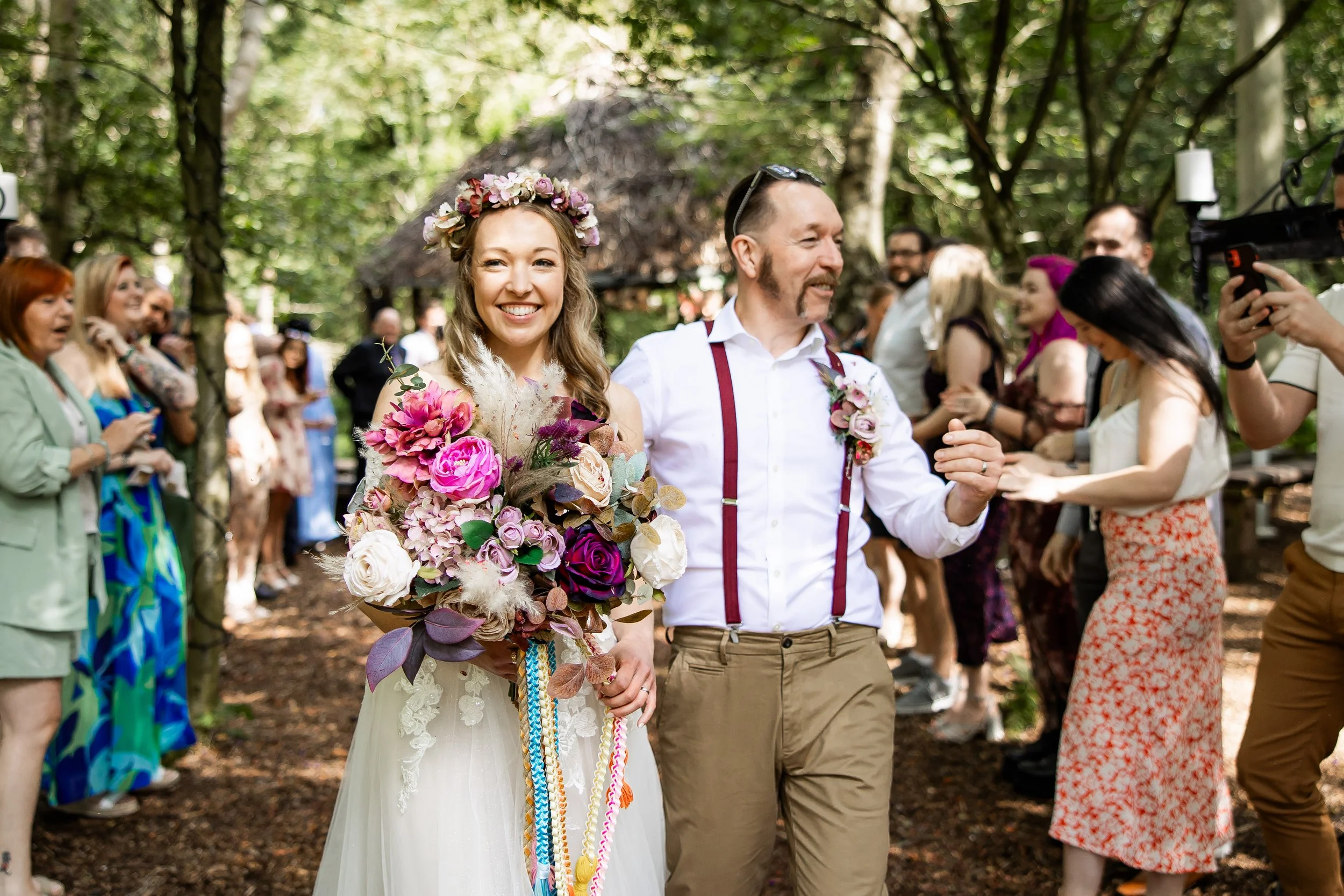 A bride with a floral crown holding a large bouquet of pink, purple, and white flowers walking outdoors with a man in suspenders during a wedding ceremony, surrounded by guests in a wooded area.