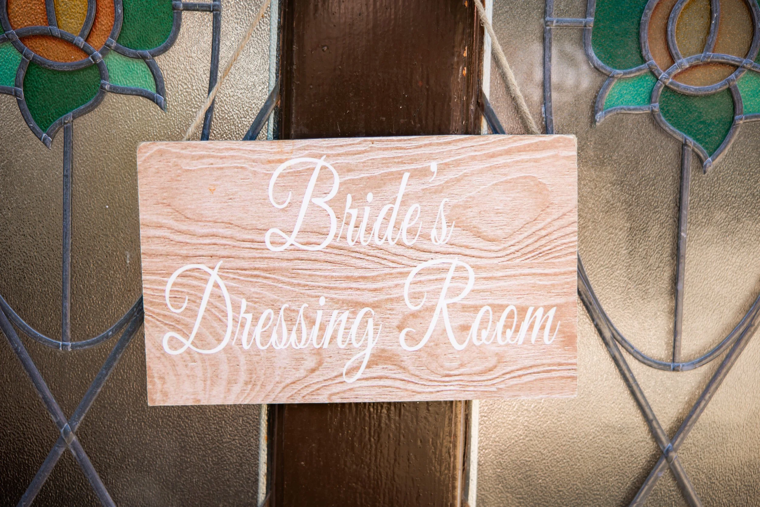 Wooden sign reading 'Brides Dressing Room' hanging on a textured glass door with decorative floral stained glass accents.
